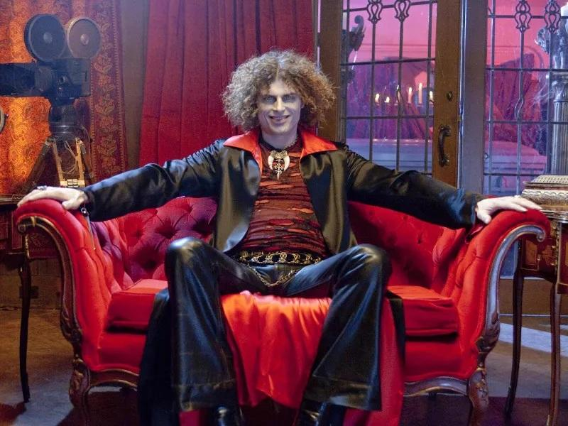 Ein lockiger Mann in Vampirkleidung sitzt breit auf einem roten Sofa, im Hintergrund eine alte Kamera und Fenster.
