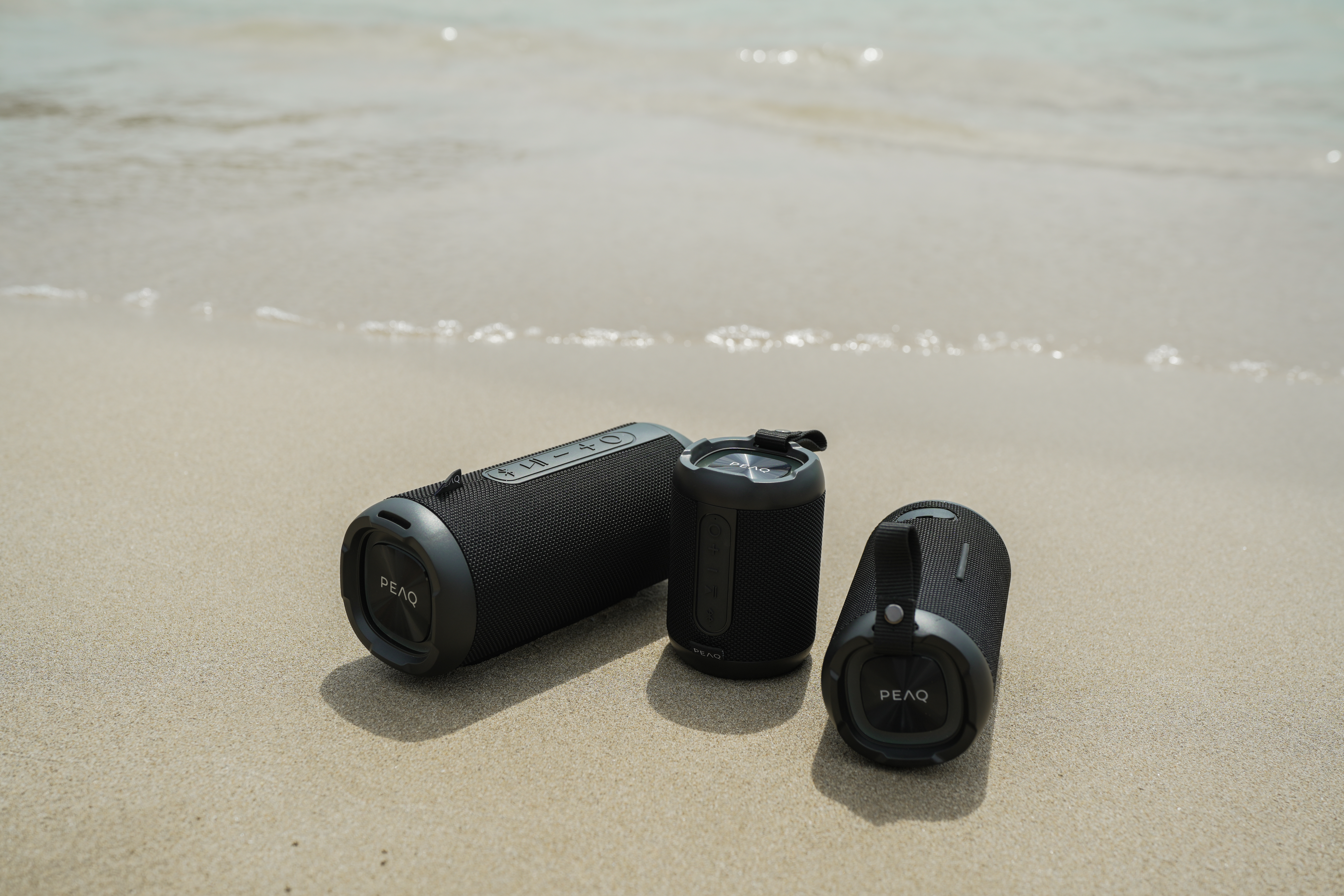 Drei schwarze PEAQ Bluetooth-Lautsprecher auf sandigem Strand am Meer.