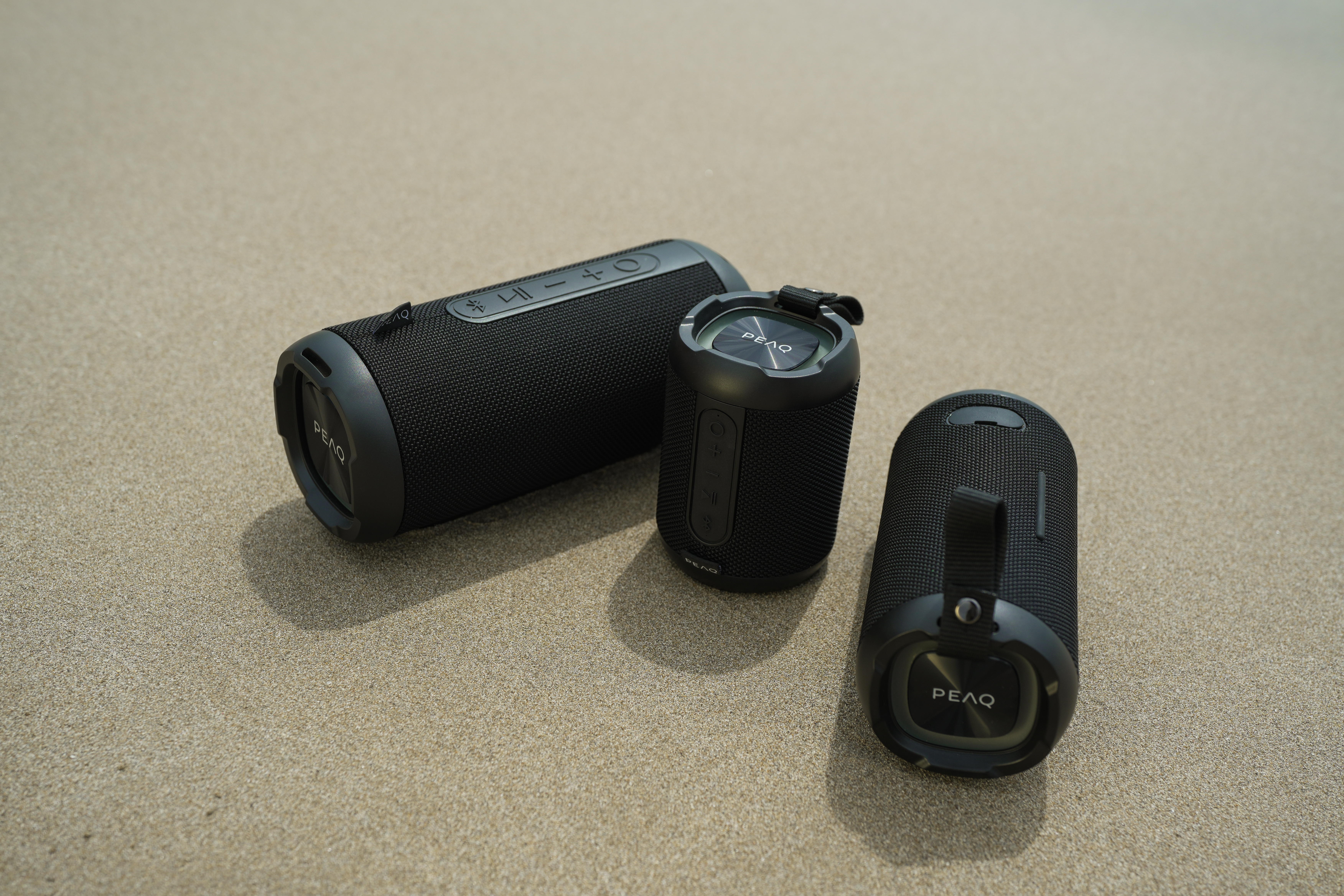 Drei schwarze PEAQ-Bluetooth-Lautsprecher liegen auf sandigem Untergrund im Freien.