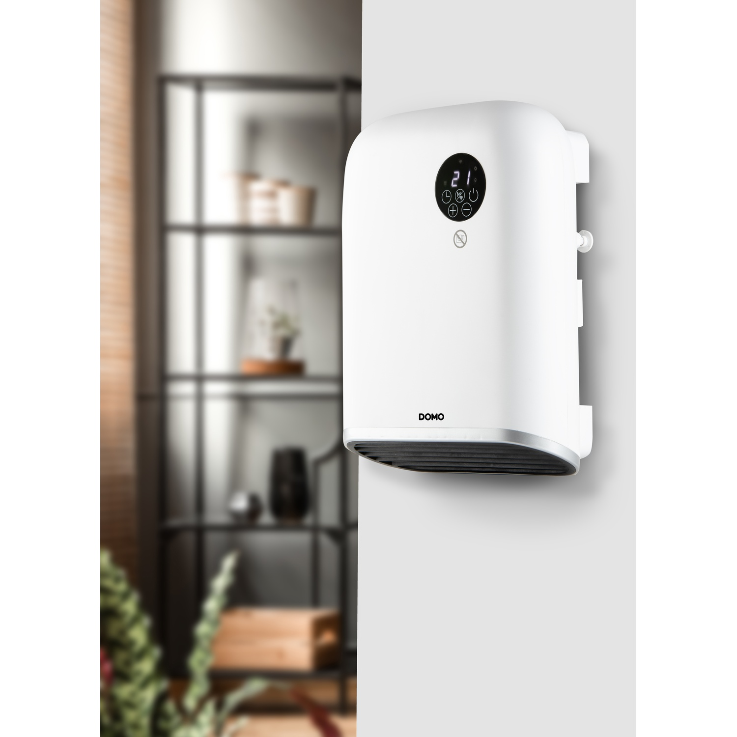 A white DOMO wall heater with a digital display mounted on a wall in a room with blurred shelving.