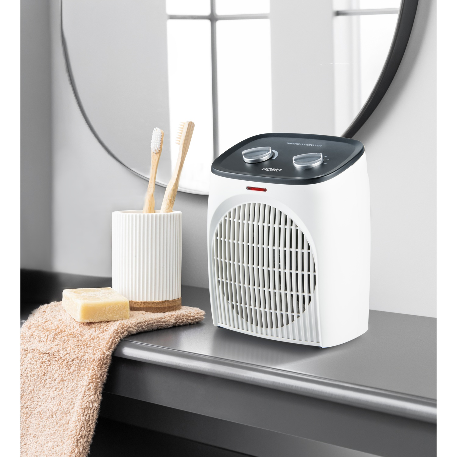 Een witte kachelventilator, tandenborstels in een houder en zeep op een handdoek voor een spiegel.