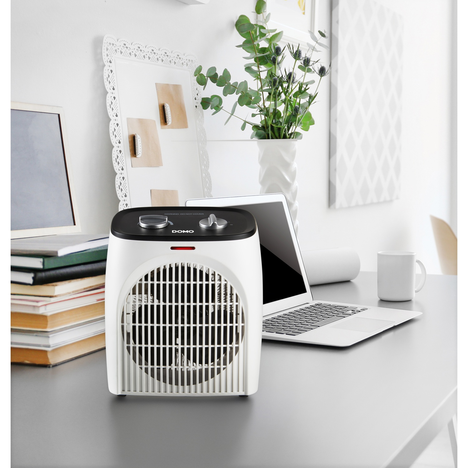Een witte Domo-ventilatorluchtkachel op een bureau met laptop, boeken en planten.