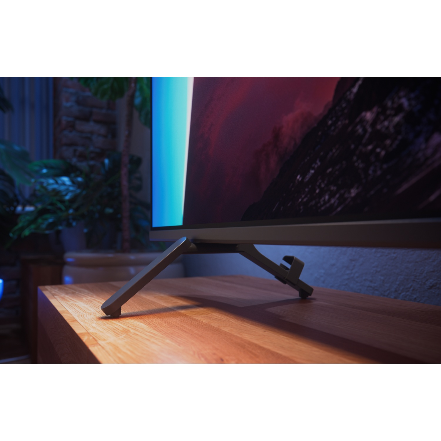 A TV with stand on a wooden table, with green plants and a wall in the background.