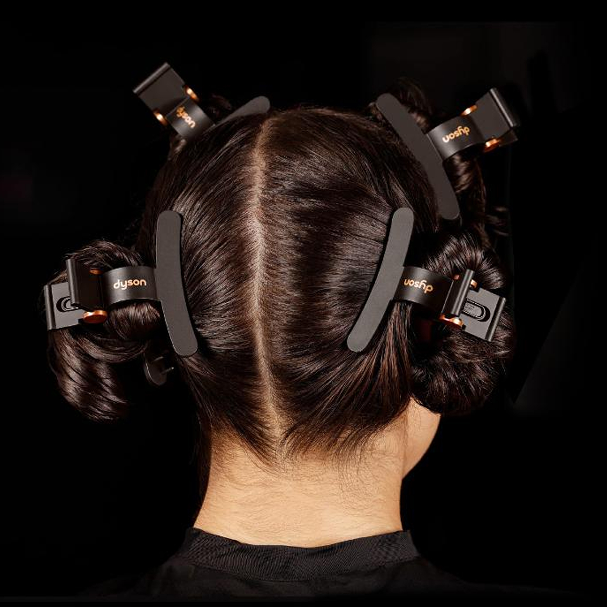Cabello oscuro, dividido y sujeto con cuatro pinzas negras Dyson sobre un fondo negro.