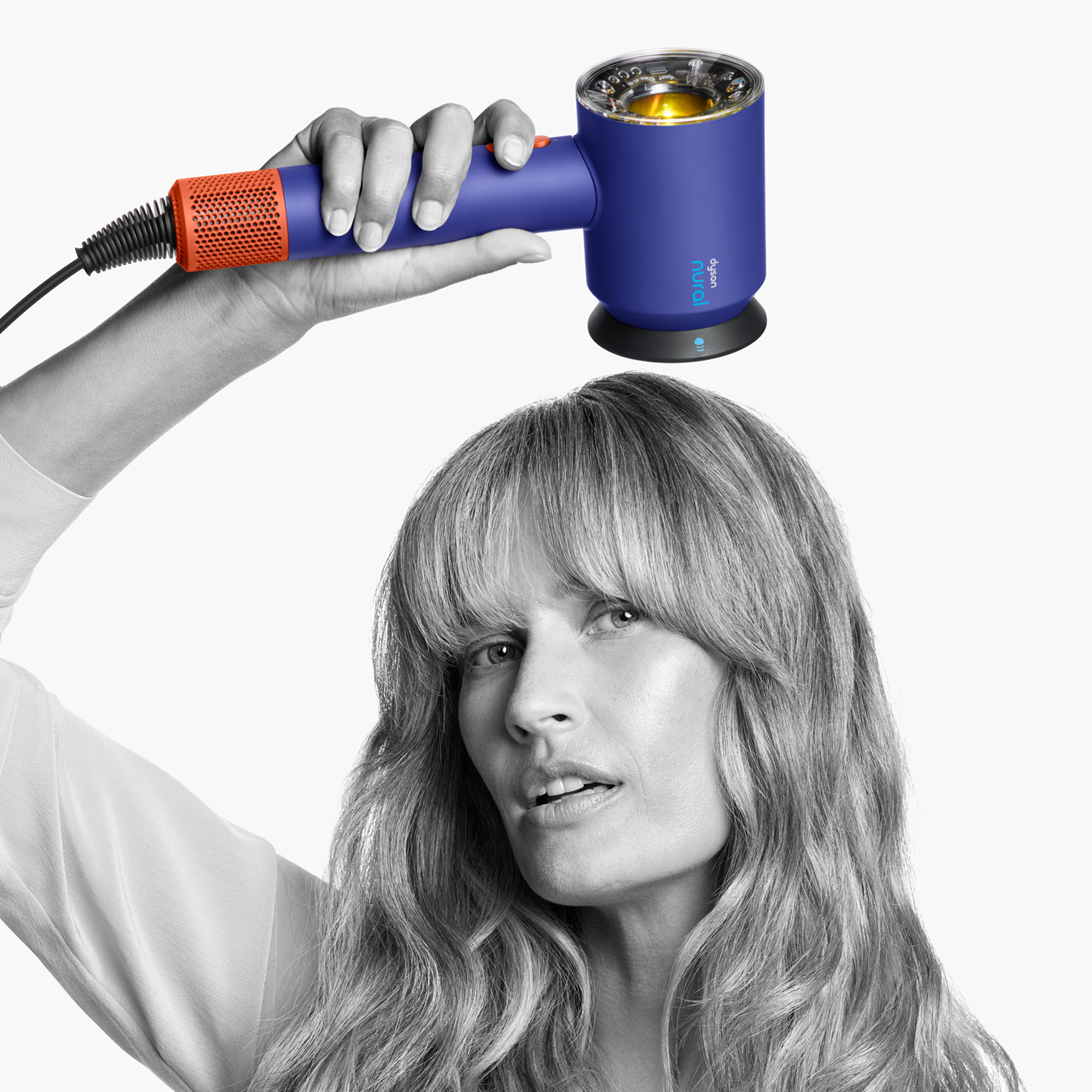 Una mujer sostiene un secador moderno morado y naranja Dyson Supersonic Nural sobre su cabeza con fondo blanco.