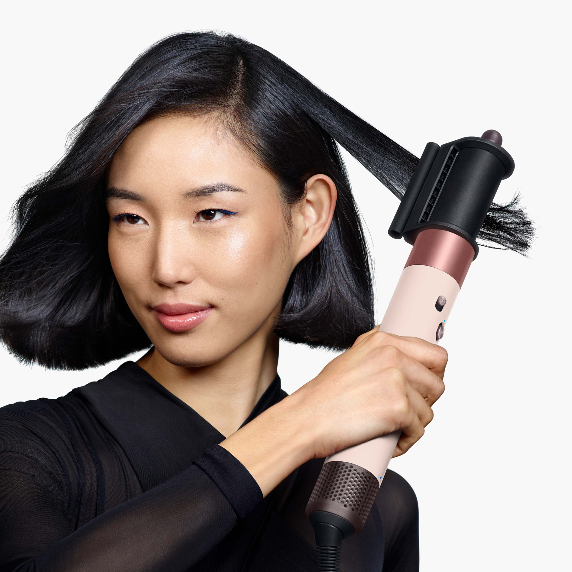 Una mujer con cabello negro corto usa una herramienta moldeadora rosa y negra en un mechón sobre fondo blanco.