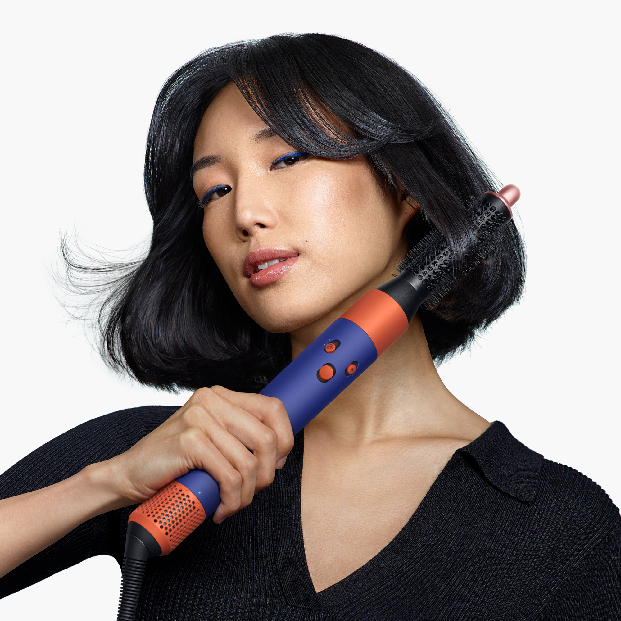 Una mujer asiática con bob negro sostiene un cepillo moldeador azul y naranja en su cabello.
