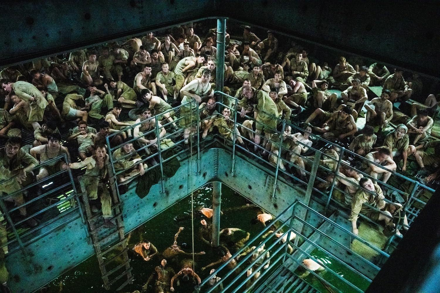Viele Männer drängen sich in einem mehrstöckigen Frachtraum mit grünem Wasser unten und blicken nach oben.