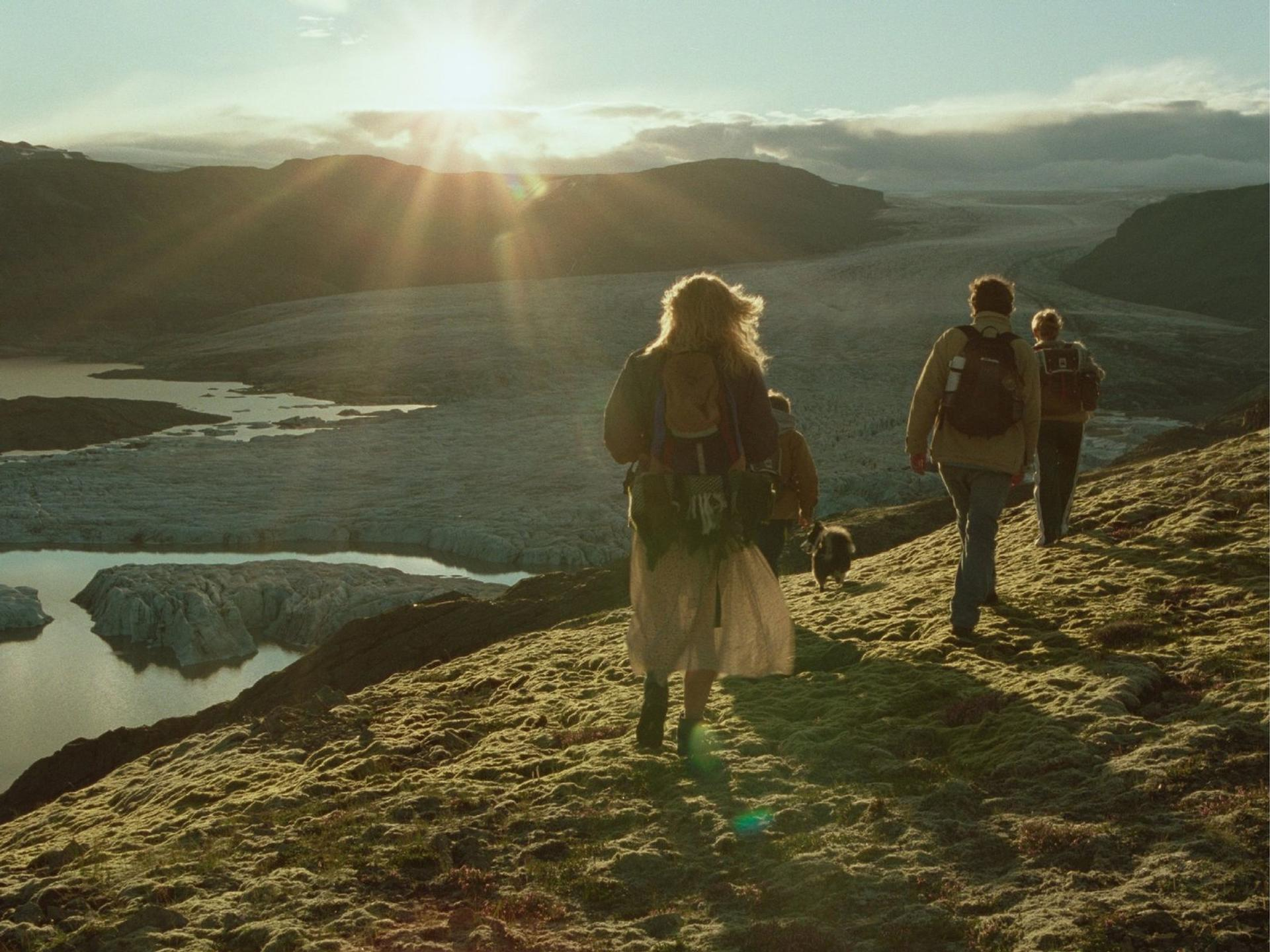 Wanderer mit Rucksäcken und Hund gehen in einer gebirgigen Landschaft mit Gletschern bei Sonnenuntergang.