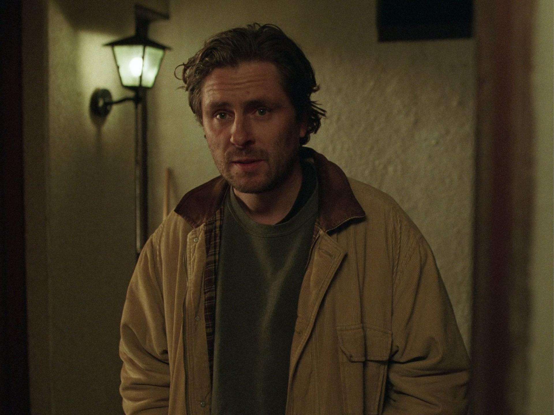 Ein Mann mit struppigem Haar trägt eine hellbraune Jacke vor einer warm erleuchteten Wandlampe.