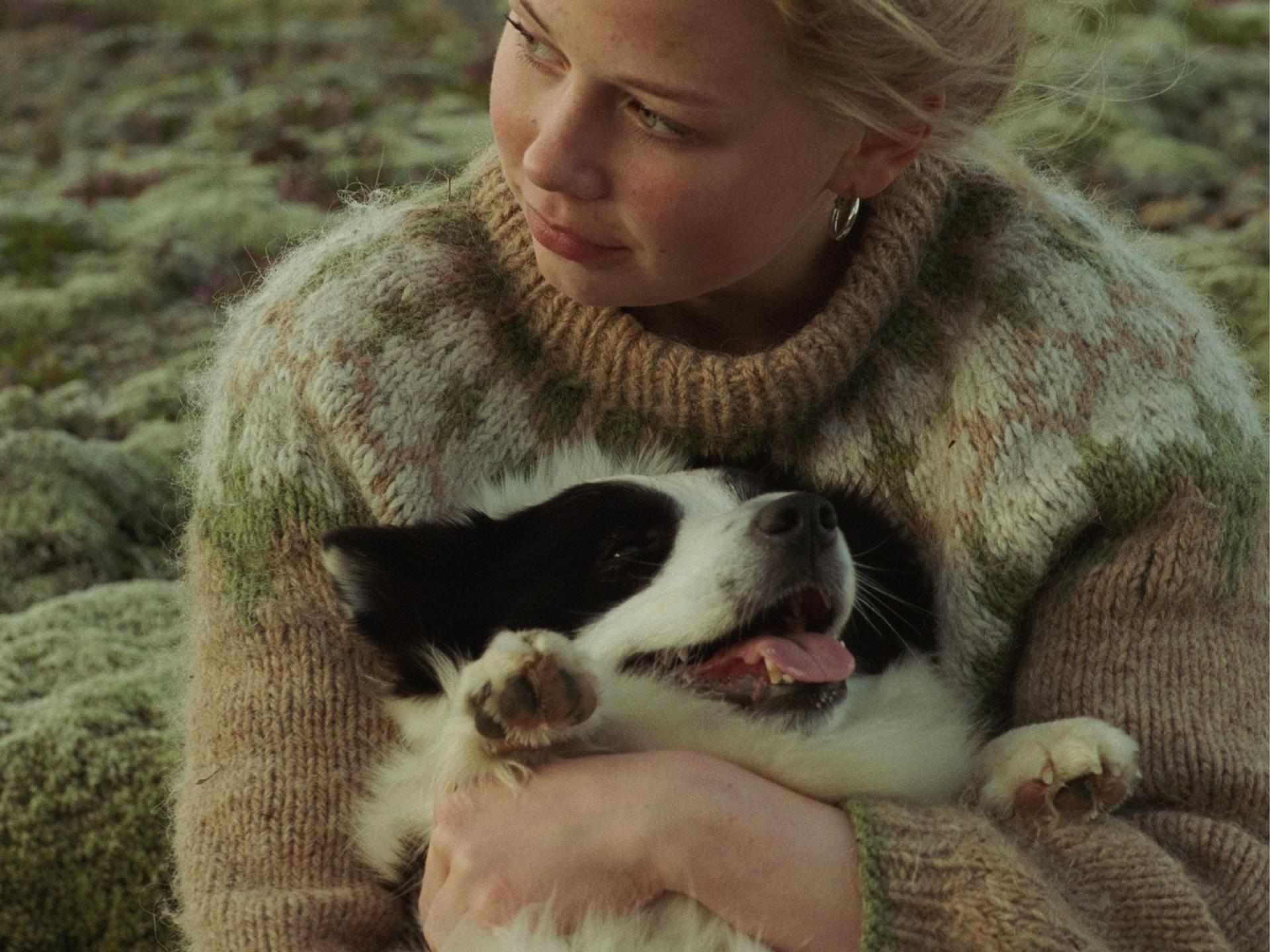 Eine blonde junge Frau in einem gemusterten Pullover hält liebevoll einen glücklichen schwarz-weißen Hund in den Armen.