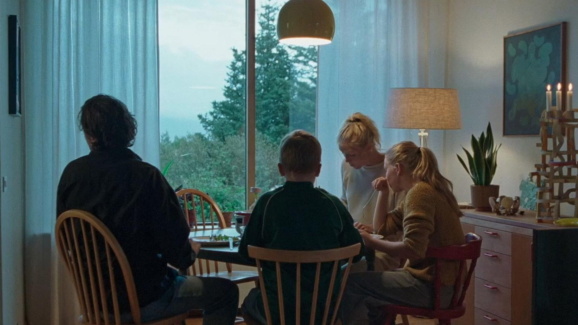 Eine vierköpfige Familie sitzt abends an einem Holztisch vor einem großen Fenster und isst in einem gemütlichen Raum.