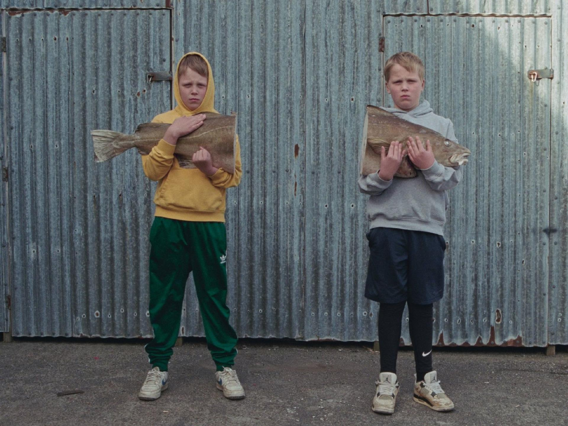 Zwei Jungen stehen vor einer Wellblechwand und halten Hälften eines großen Fisches.