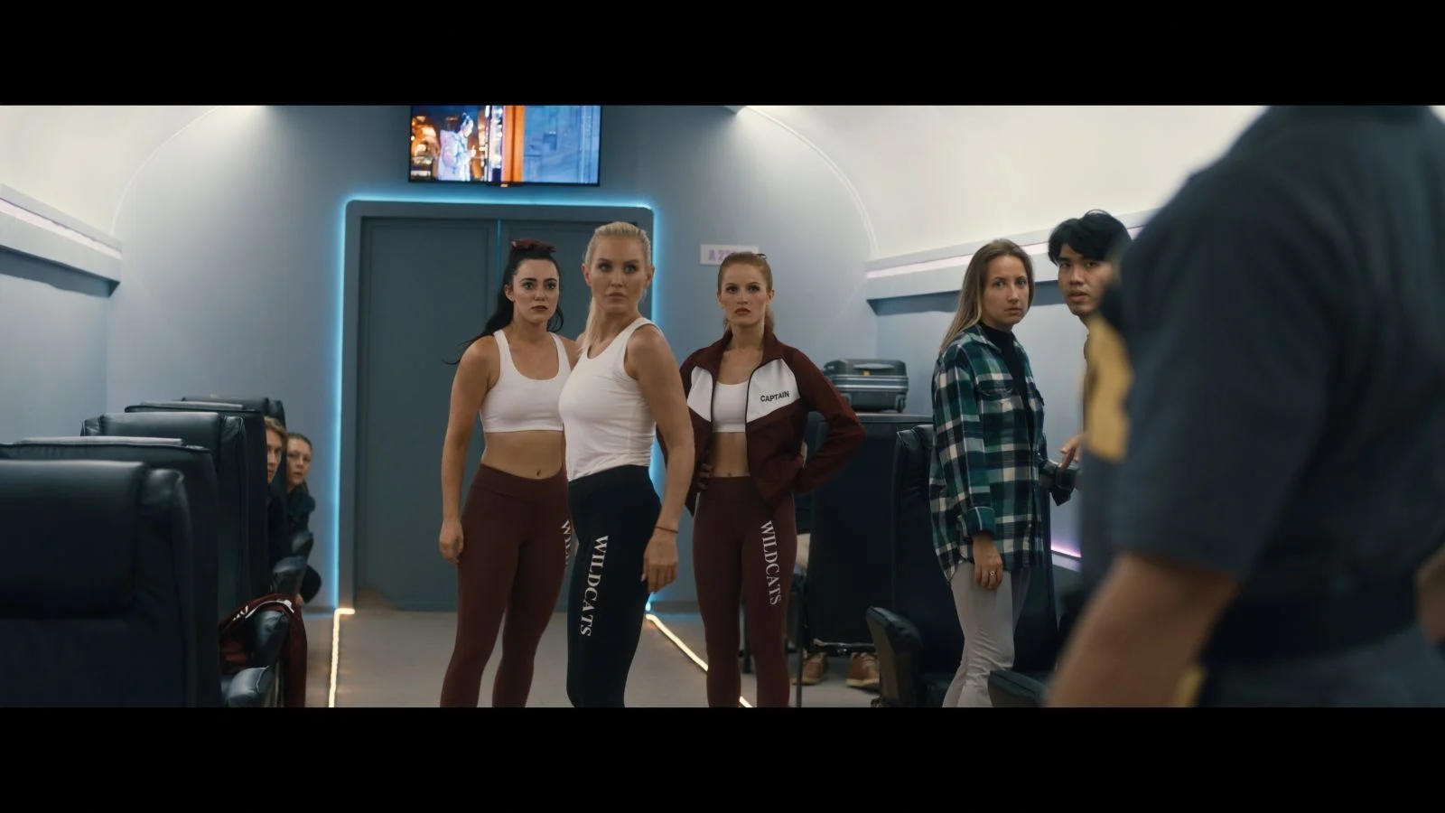 Eine Gruppe von Menschen in Sportbekleidung steht in einer futuristischen Kabine und blickt zu einer Person.