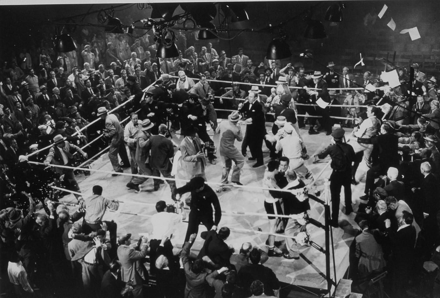 Eine Menschenmenge drängt sich in und um einen Boxring, während Scheinwerfer die Szene erhellen.