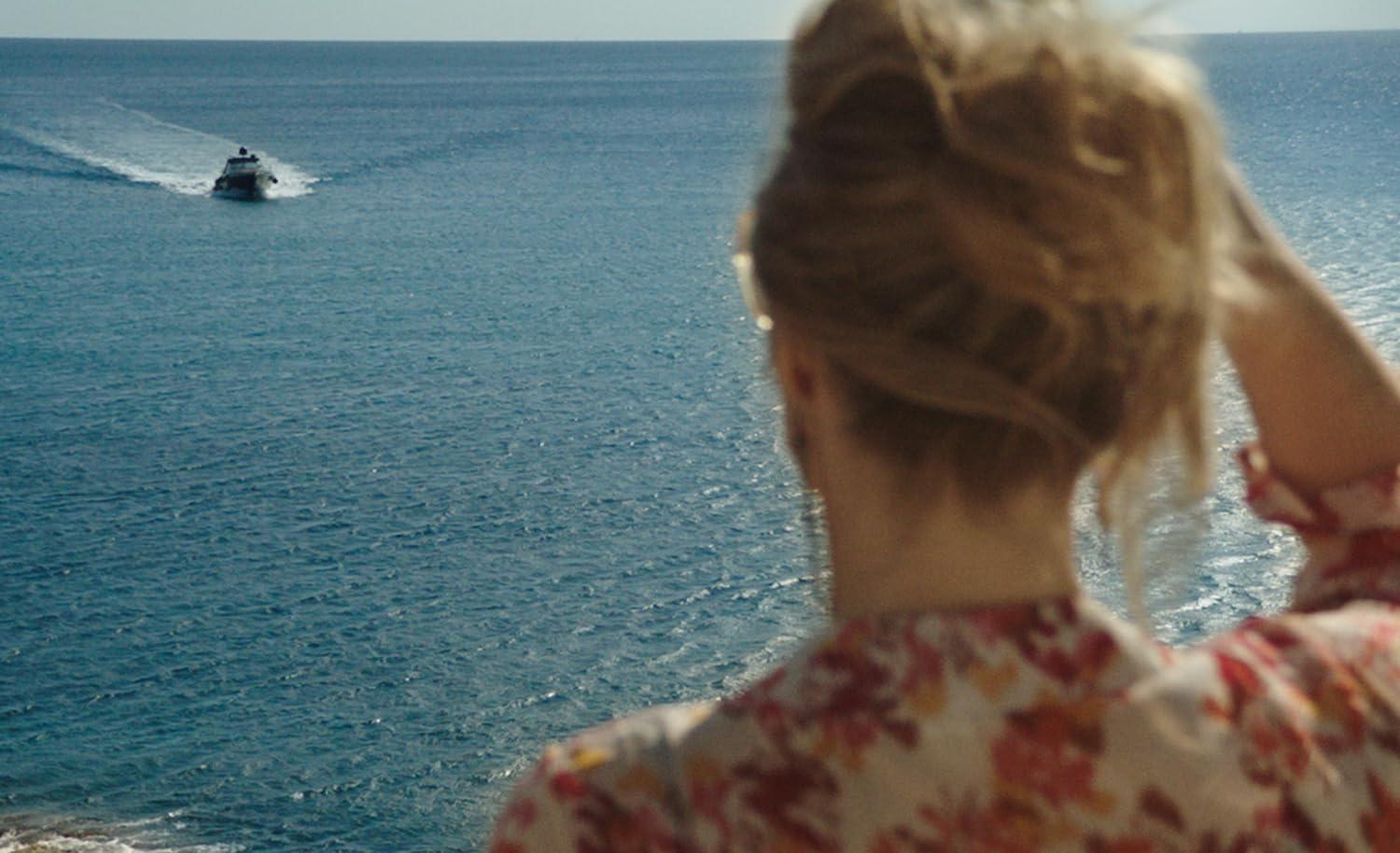Eine Frau in bunter Bluse schaut von oben auf das blaue Meer, auf dem ein kleines Boot fährt.