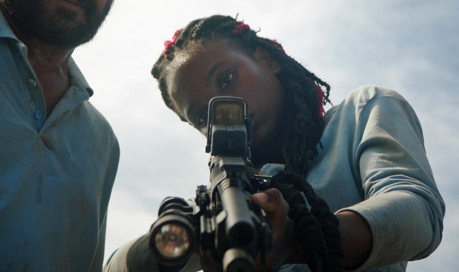 Eine Frau mit Rastafari-Frisur zielt entschlossen mit einem Gewehr nach vorne, während ein Mann daneben steht.
