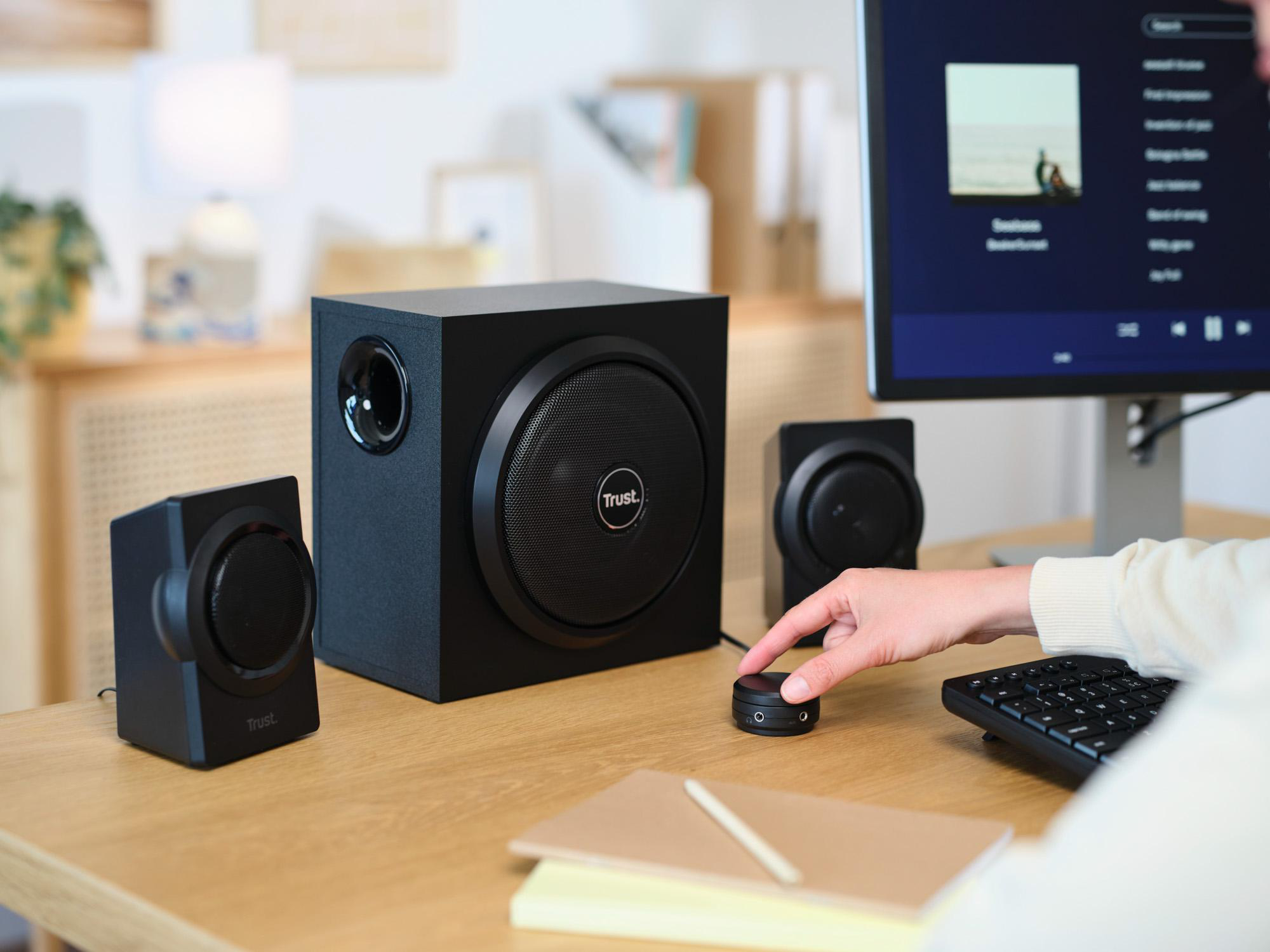 Eine Person reguliert die Lautstärke neben schwarzen Trust-Lautsprechern und einem PC auf einem Holztisch.