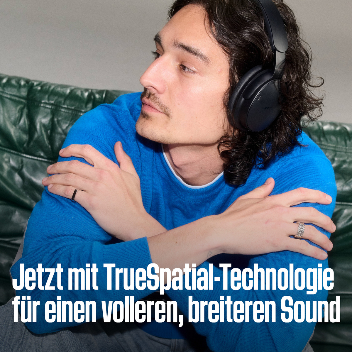 Ein Mann mit dunklen Haaren und blauem Pullover trägt schwarze Kopfhörer auf einer grünen Couch.
