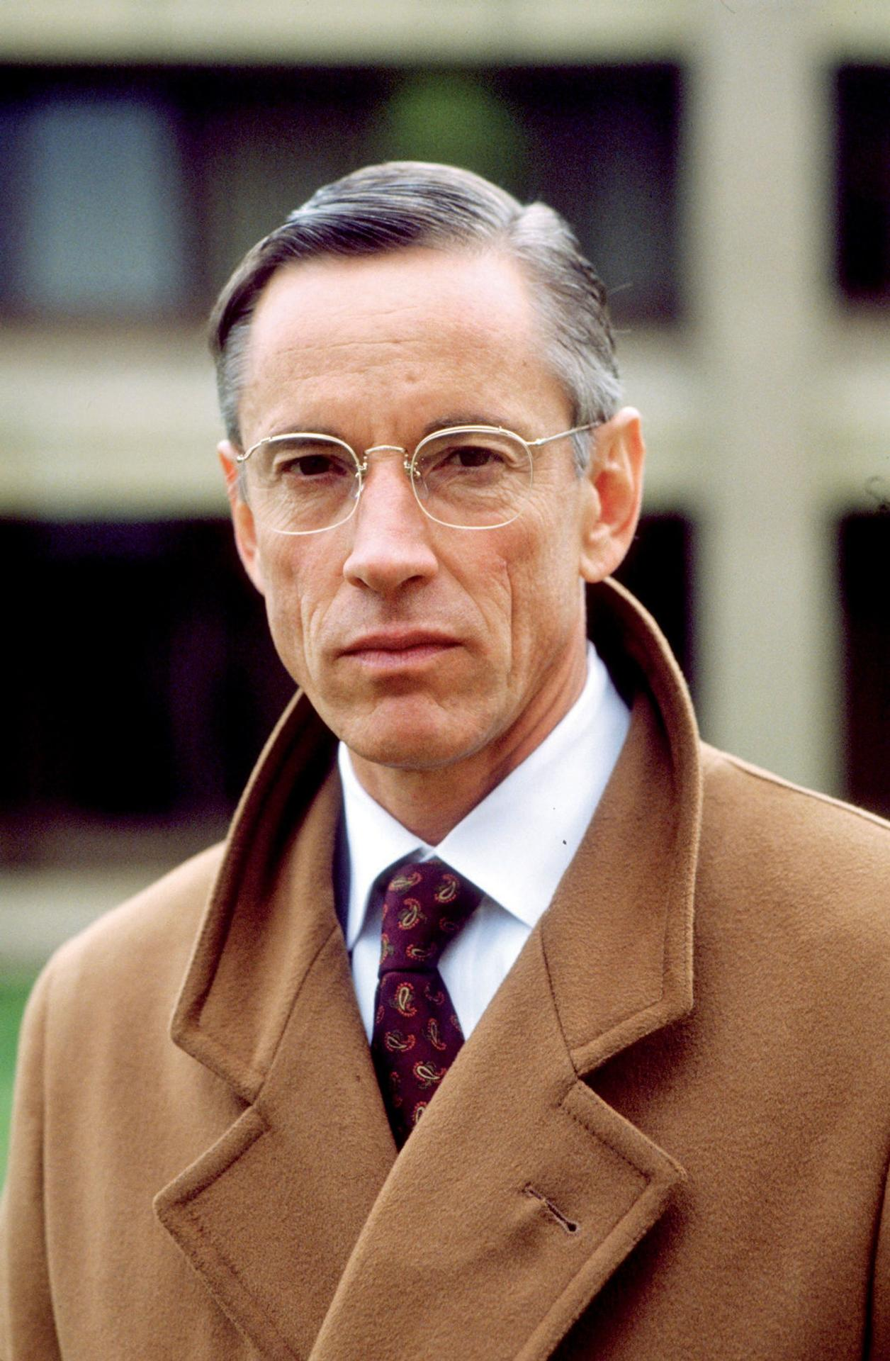 Ein Mann mit Brille trägt einen braunen Mantel und eine gemusterte Krawatte vor einem unscharfen Hintergrund.