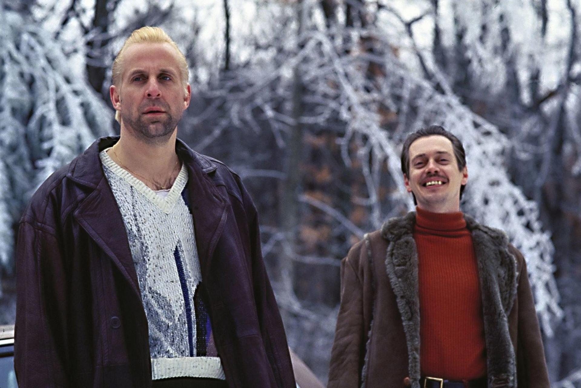 Zwei Männer stehen draußen im Schnee; der linke trägt eine Lederjacke, der rechte einen roten Rolkragenpullover.