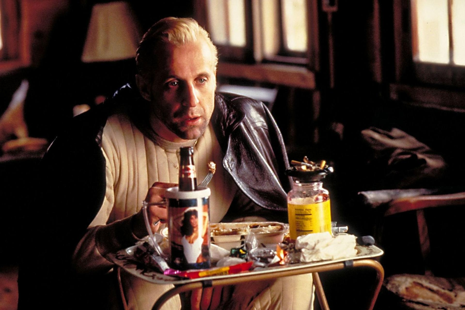 Ein blondhaariger Mann sitzt vor einem Tisch mit Lebensmitteln und Getränken in einem Raum.