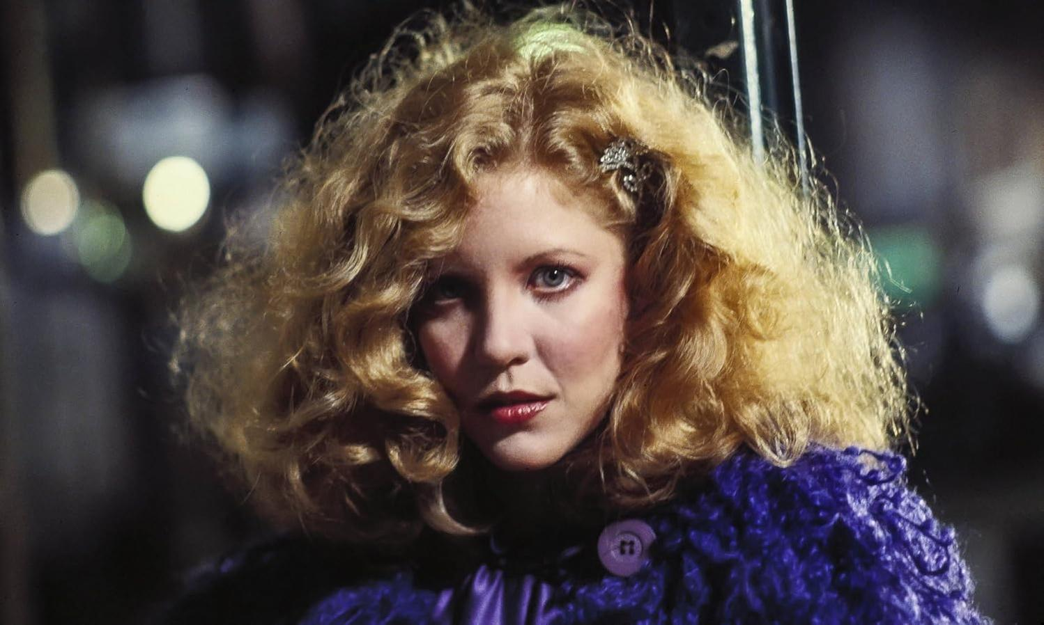 Eine blonde Frau mit lockigem Haar und violettem Mantel blickt ernst in die Kamera.