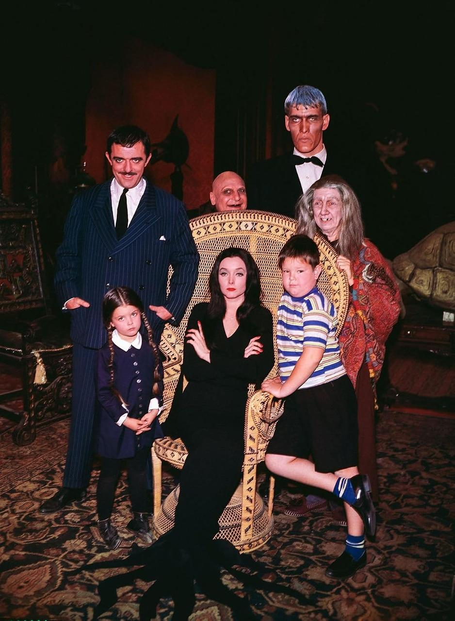 Die Addams Family posiert gemeinsam in einem dunklen Raum, die Mutter sitzt in einem großen Korbstuhl.