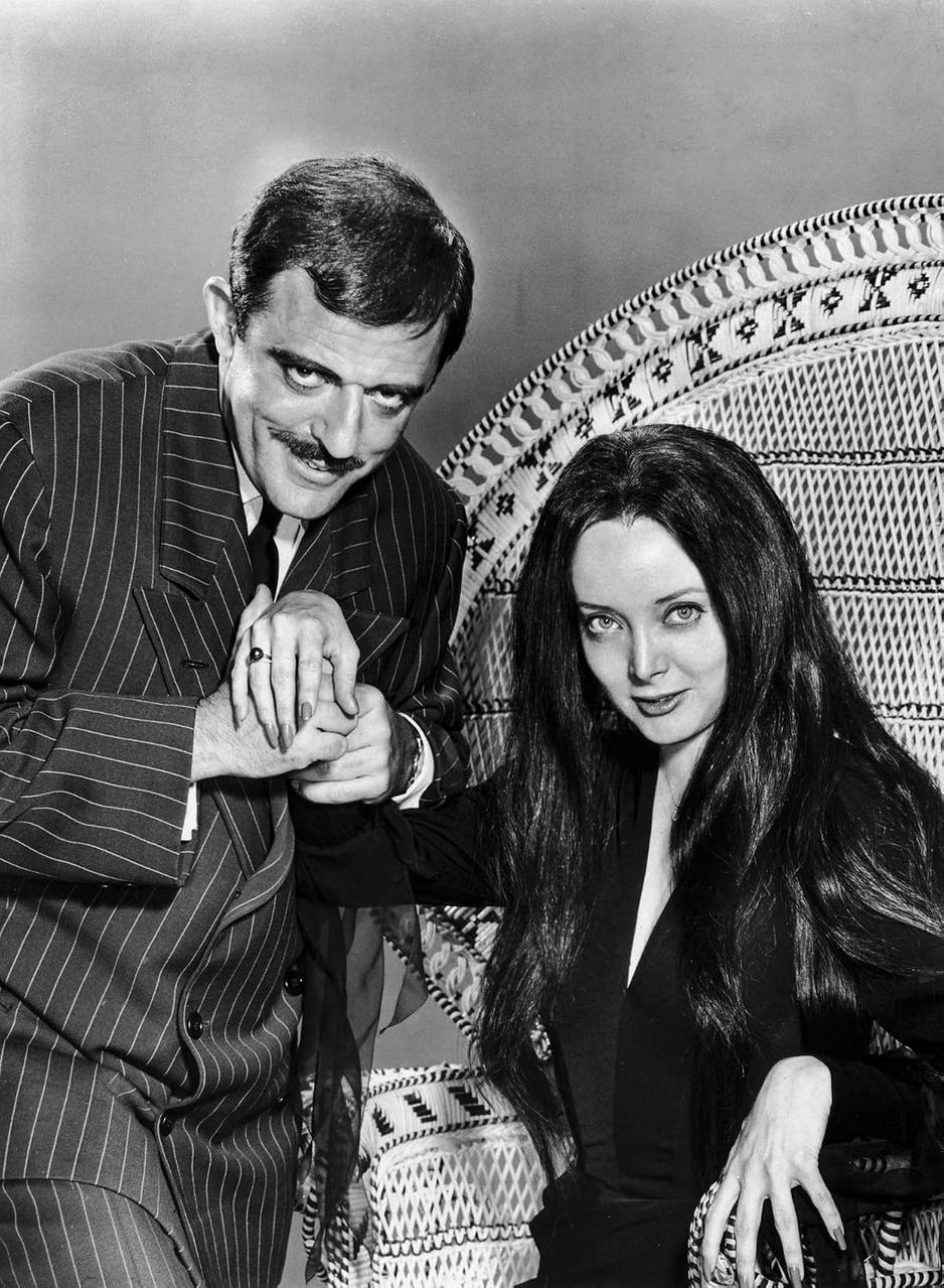 Ein schwarz-weißes Bild von Gomez und Morticia Addams in einem Flechtsessel, Gomez küsst ihre Hand.