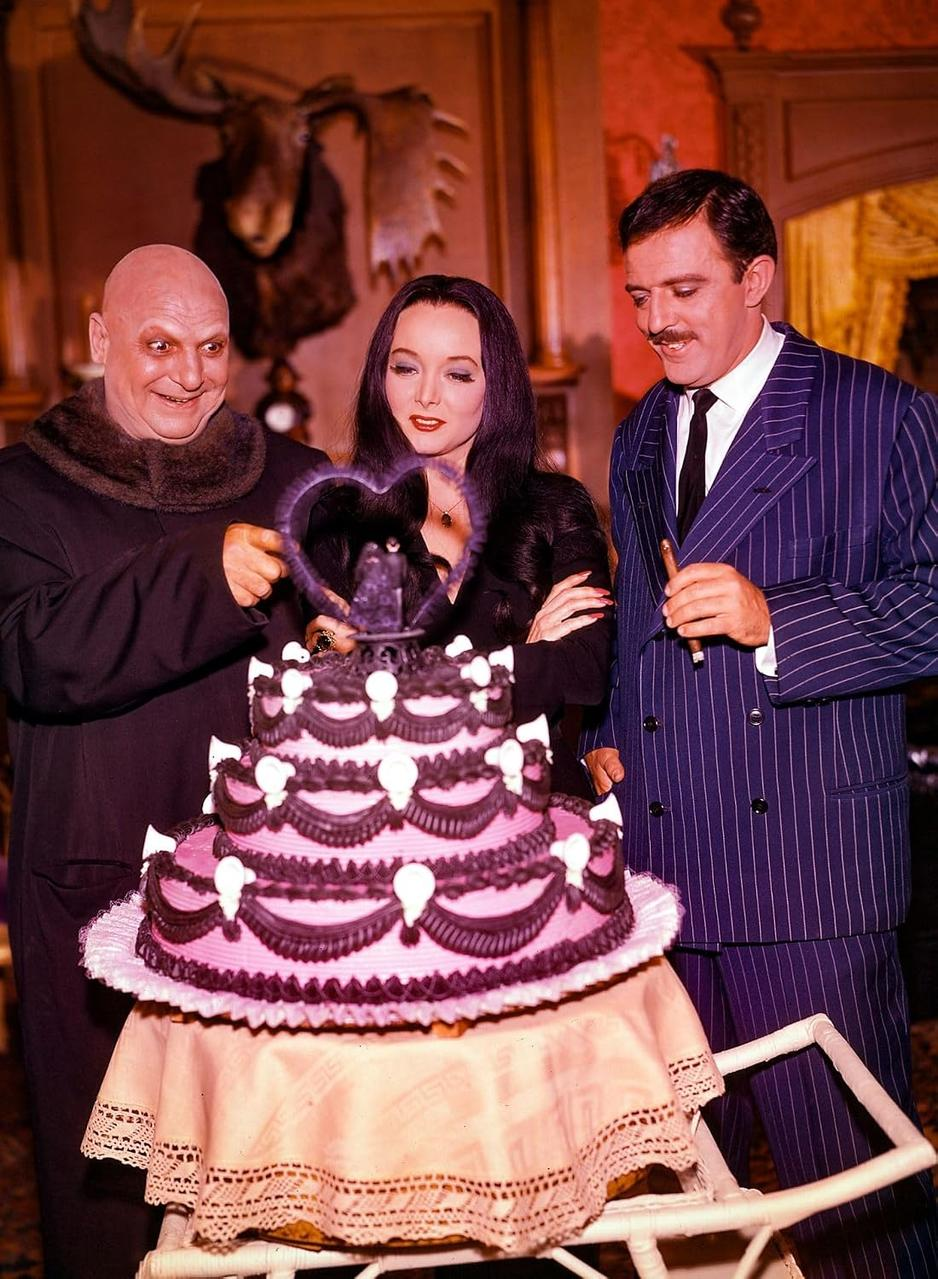 Drei Figuren stehen um eine grosse rosafarbene und schwarze Torte mit einer Herzdekoration.