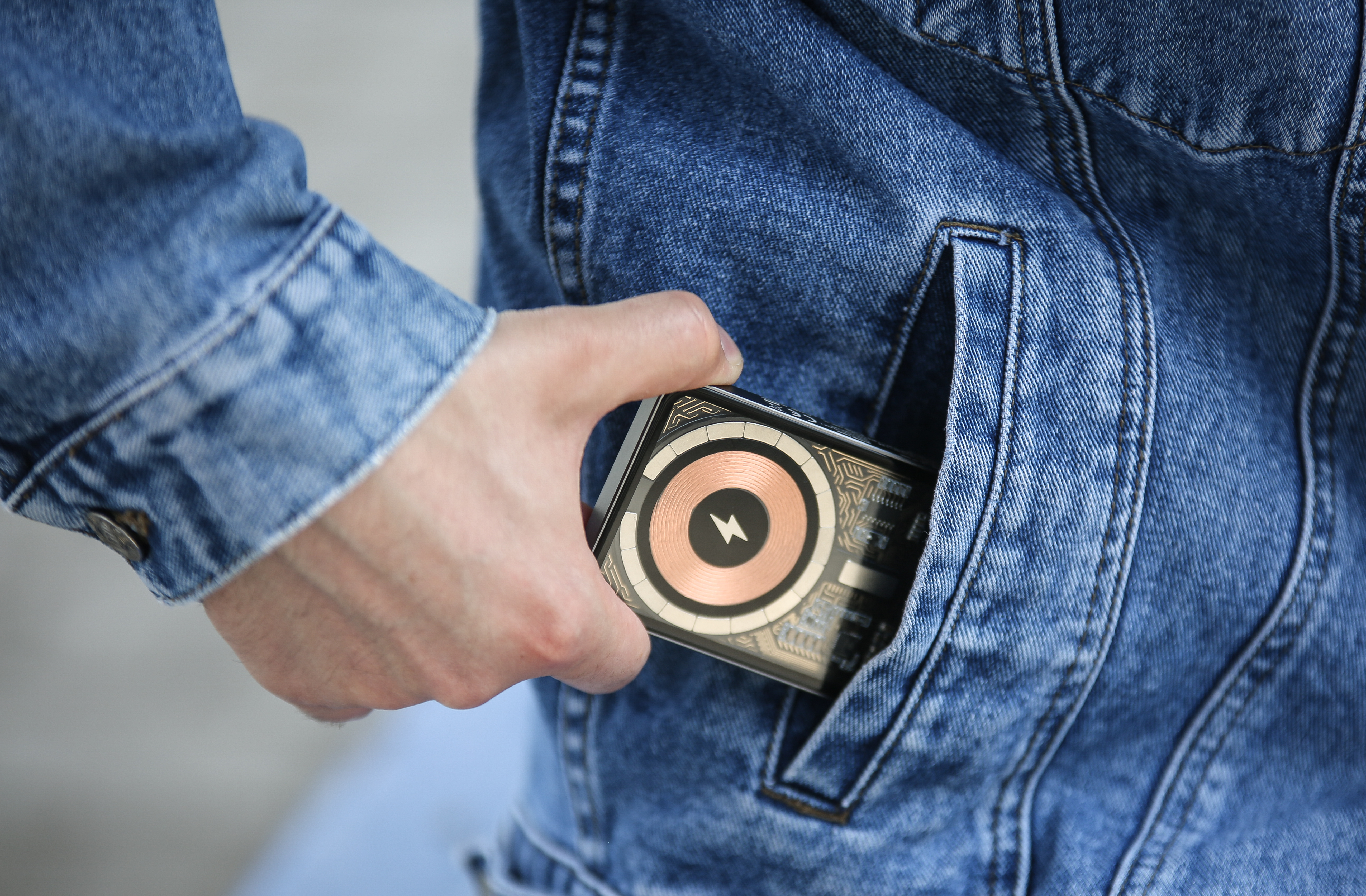 Eine Hand steckt eine Powerbank mit kupferfarbenem Spulendesign in die Tasche einer blauen Jeansjacke.