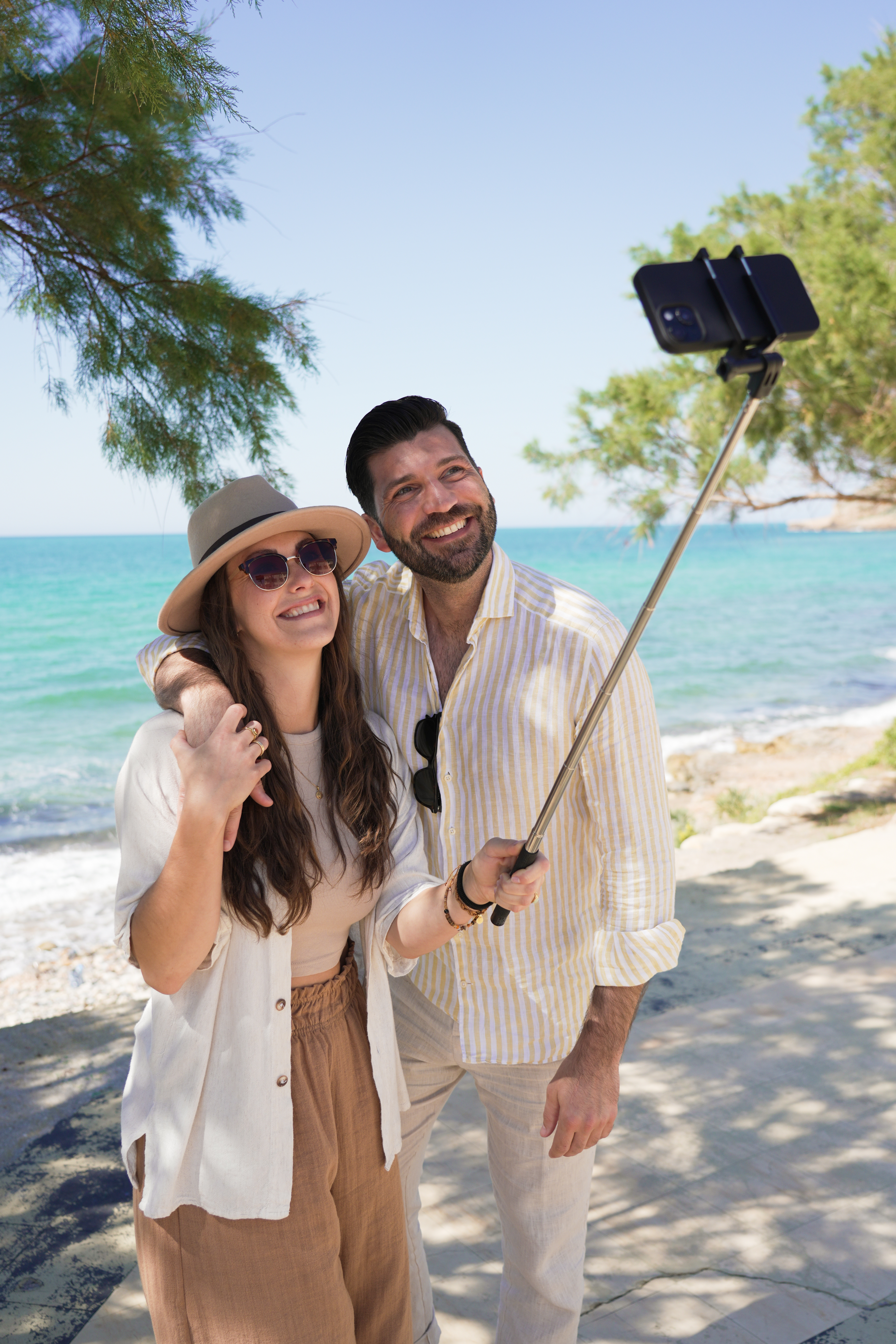 Paar macht ein Selfie mit Selfie-Stick am sonnigen Strand mit blauem Meer.