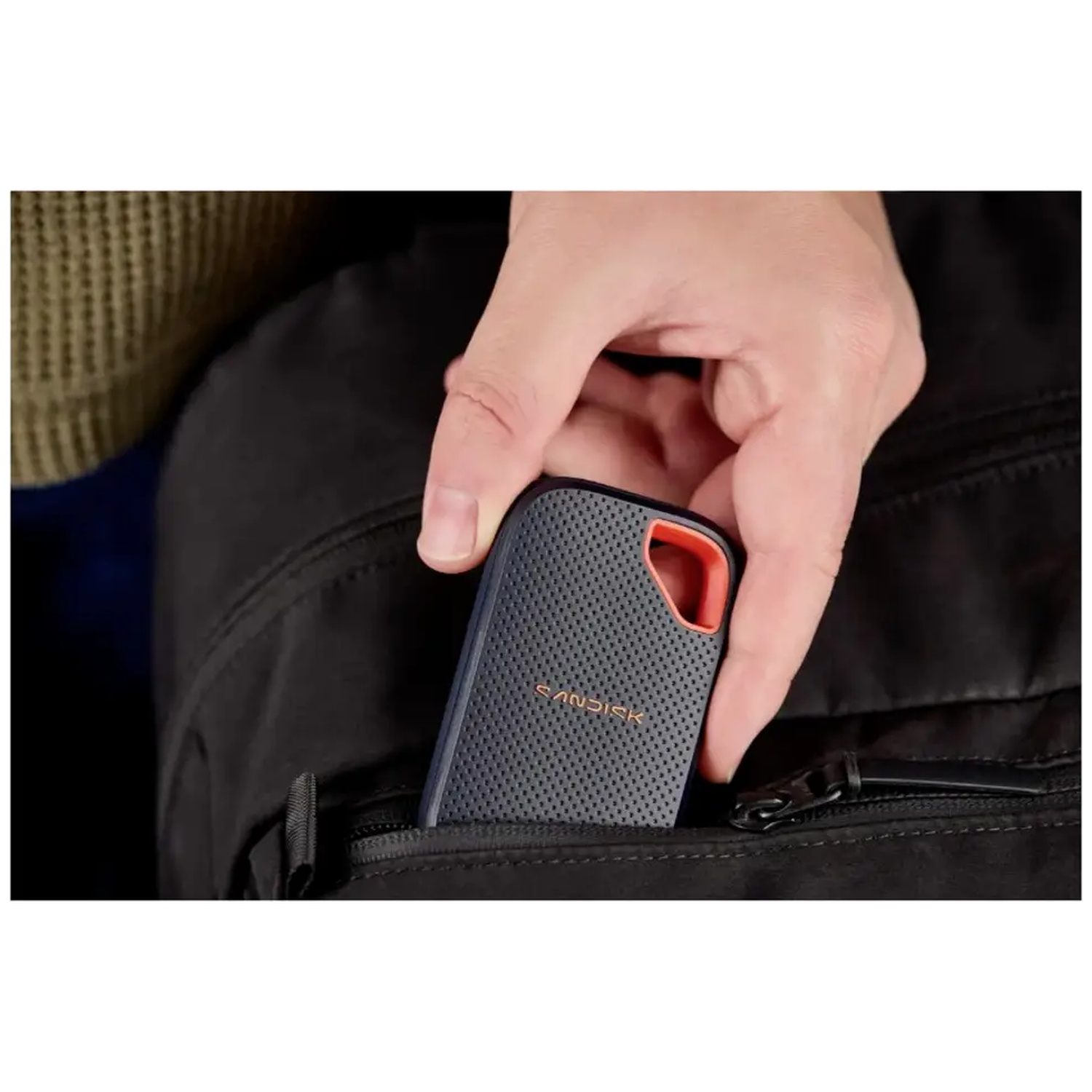Une main place un SSD SanDisk texturé noir dans la poche d'un sac à dos noir.