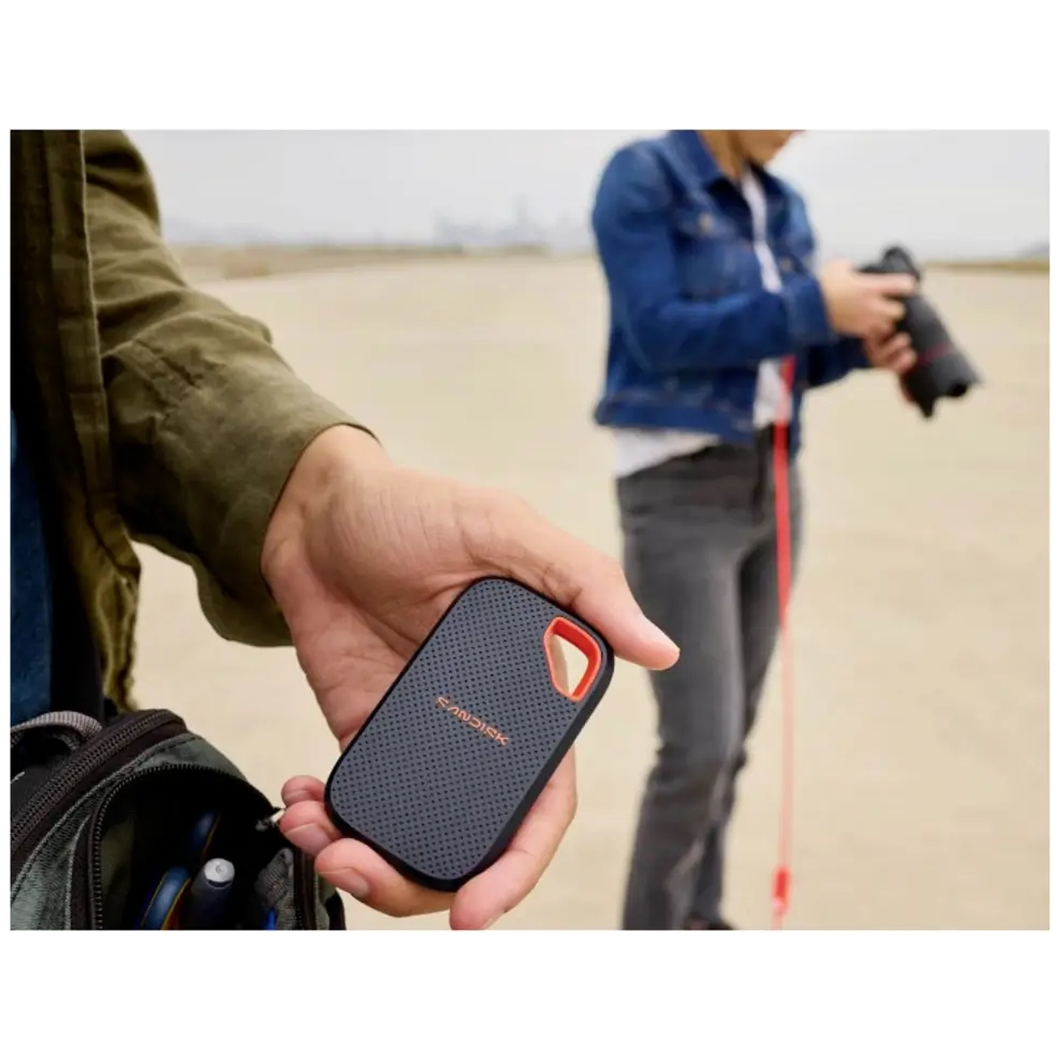 Une personne tient un petit disque dur SanDisk noir dans sa main tandis qu'une autre prend des photos en arrière-plan.