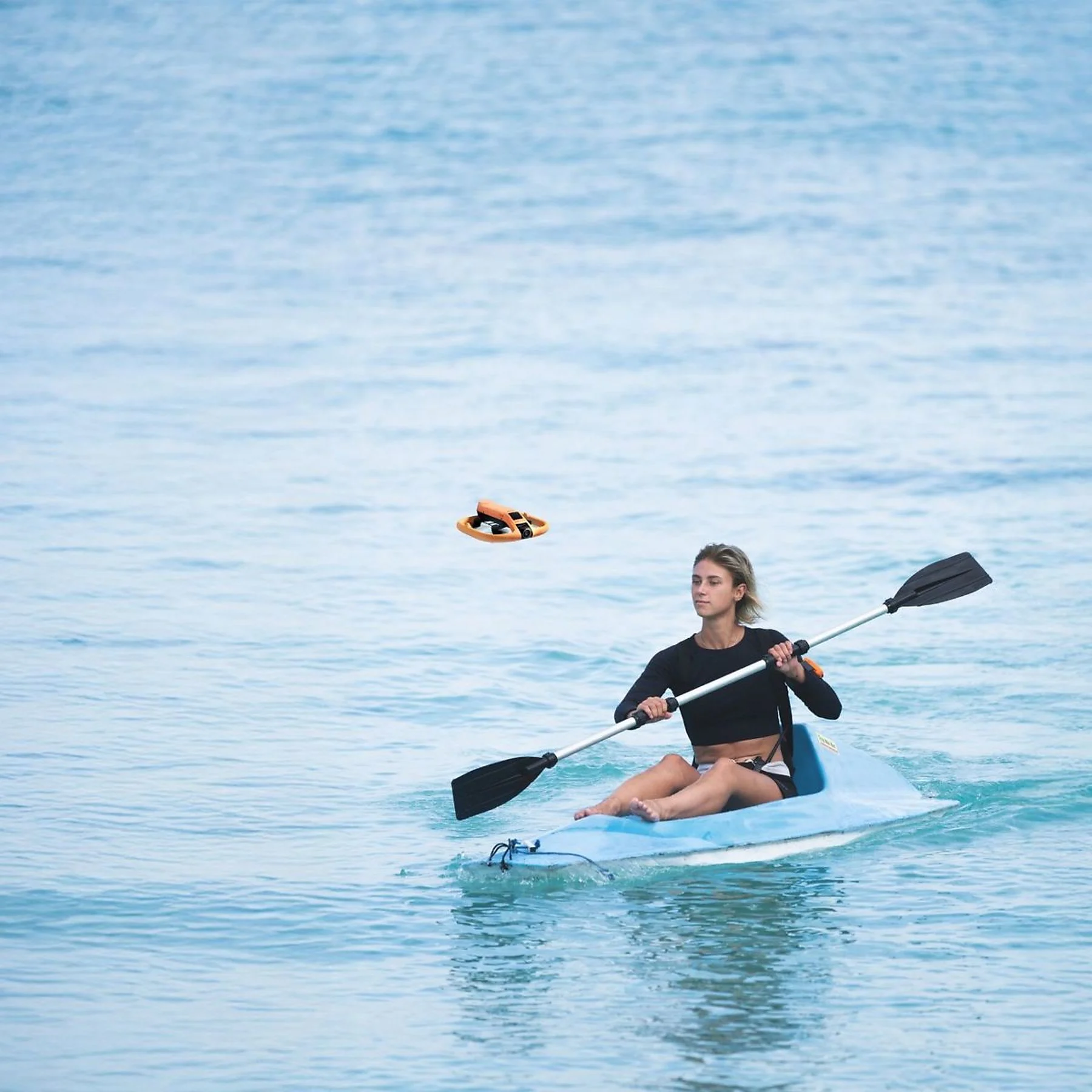 Una mujer rema en un kayak azul en aguas tranquilas del mar mientras un objeto amarillo vuela en el aire.