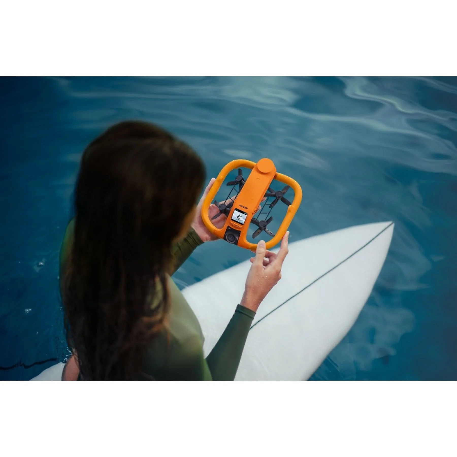 Una mujer en traje de neopreno verde sostiene un dron naranja mientras está en una tabla de surf sobre agua azul.