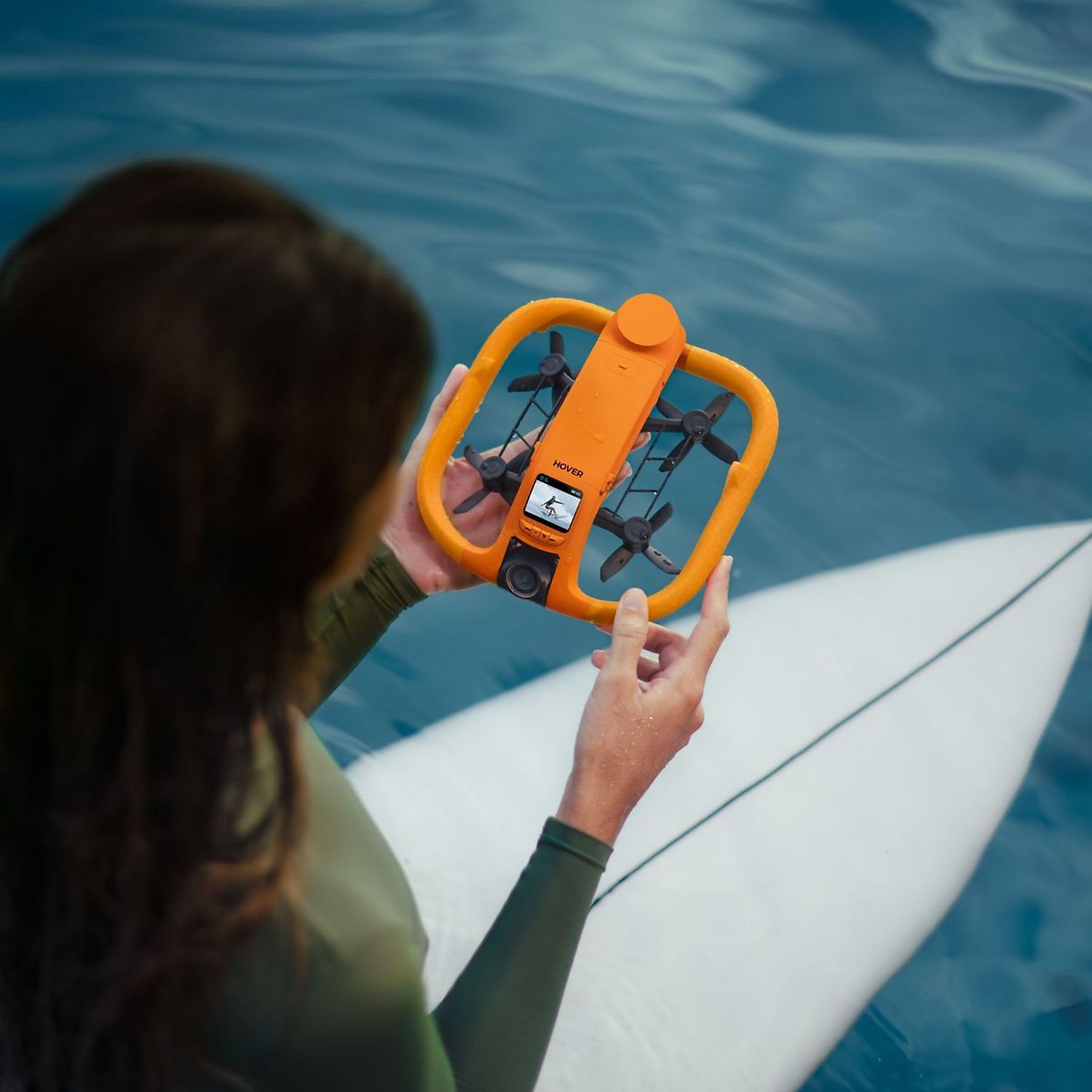 Una mujer sostiene un dron naranja sobre una tabla de surf en agua azul.