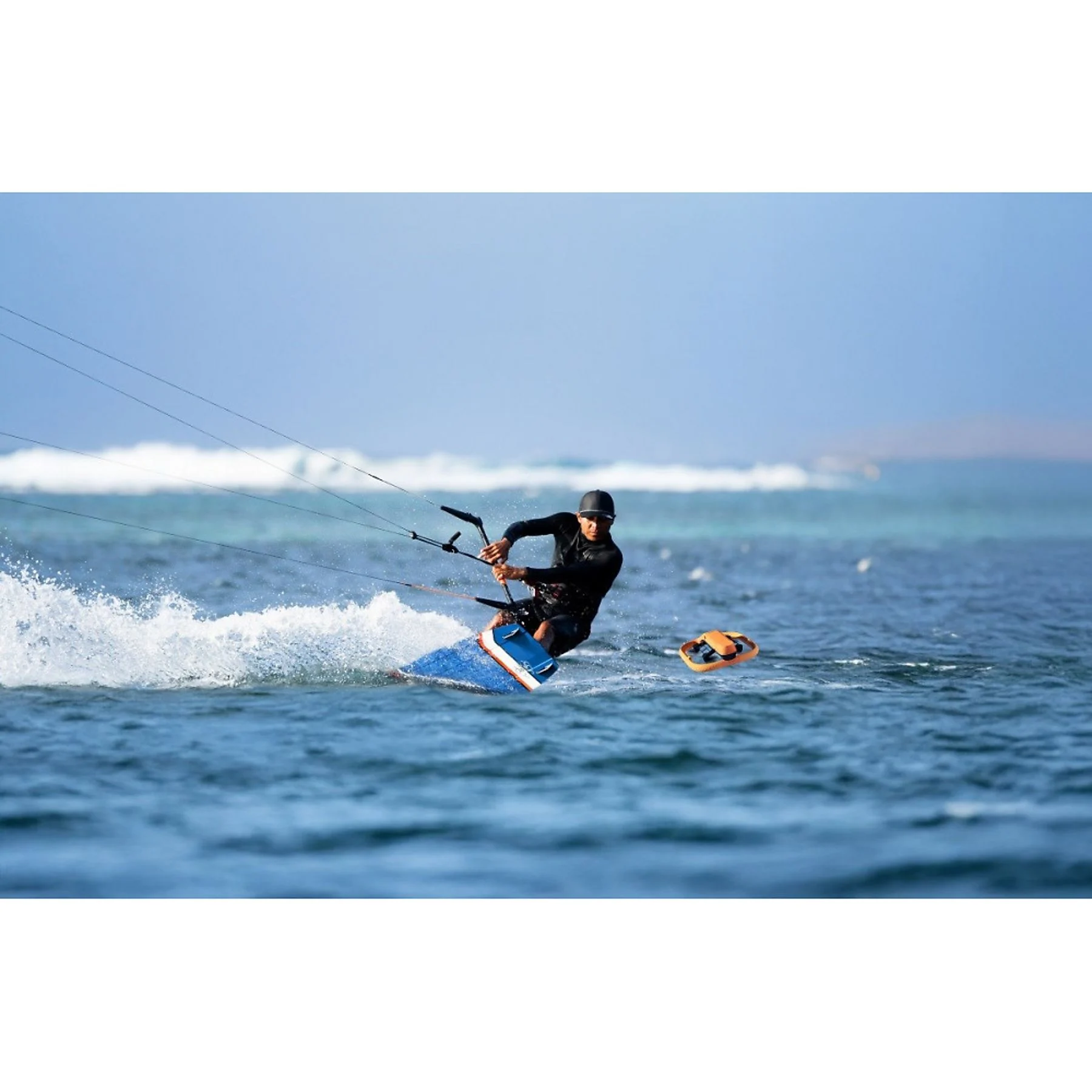 Un kitesurfista con ropa negra navega en una tabla azul por el mar azul, creando salpicaduras de agua.