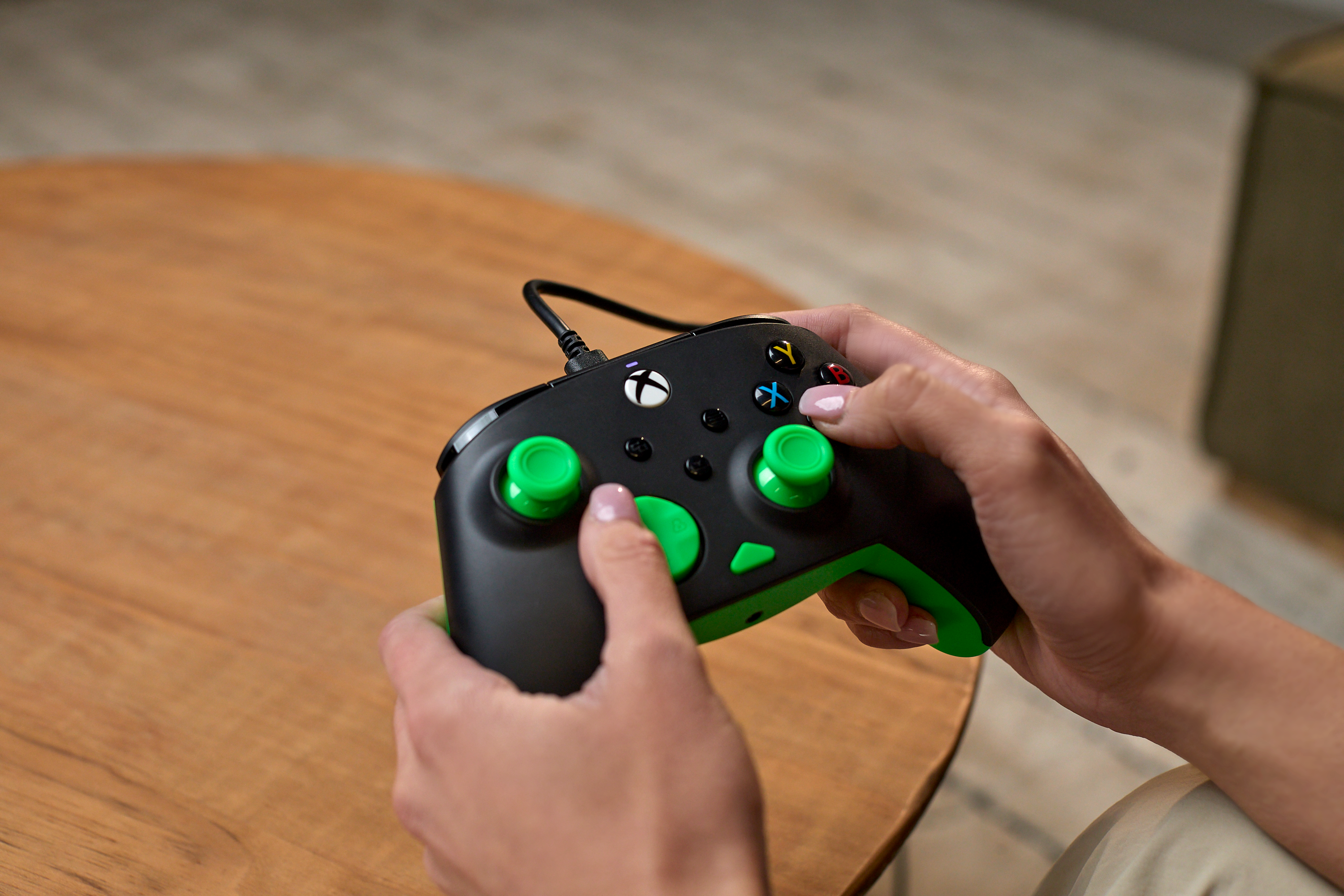 Osoba trzymająca czarny kontroler Xbox z zielonymi akcentami przed drewnianym stółem.