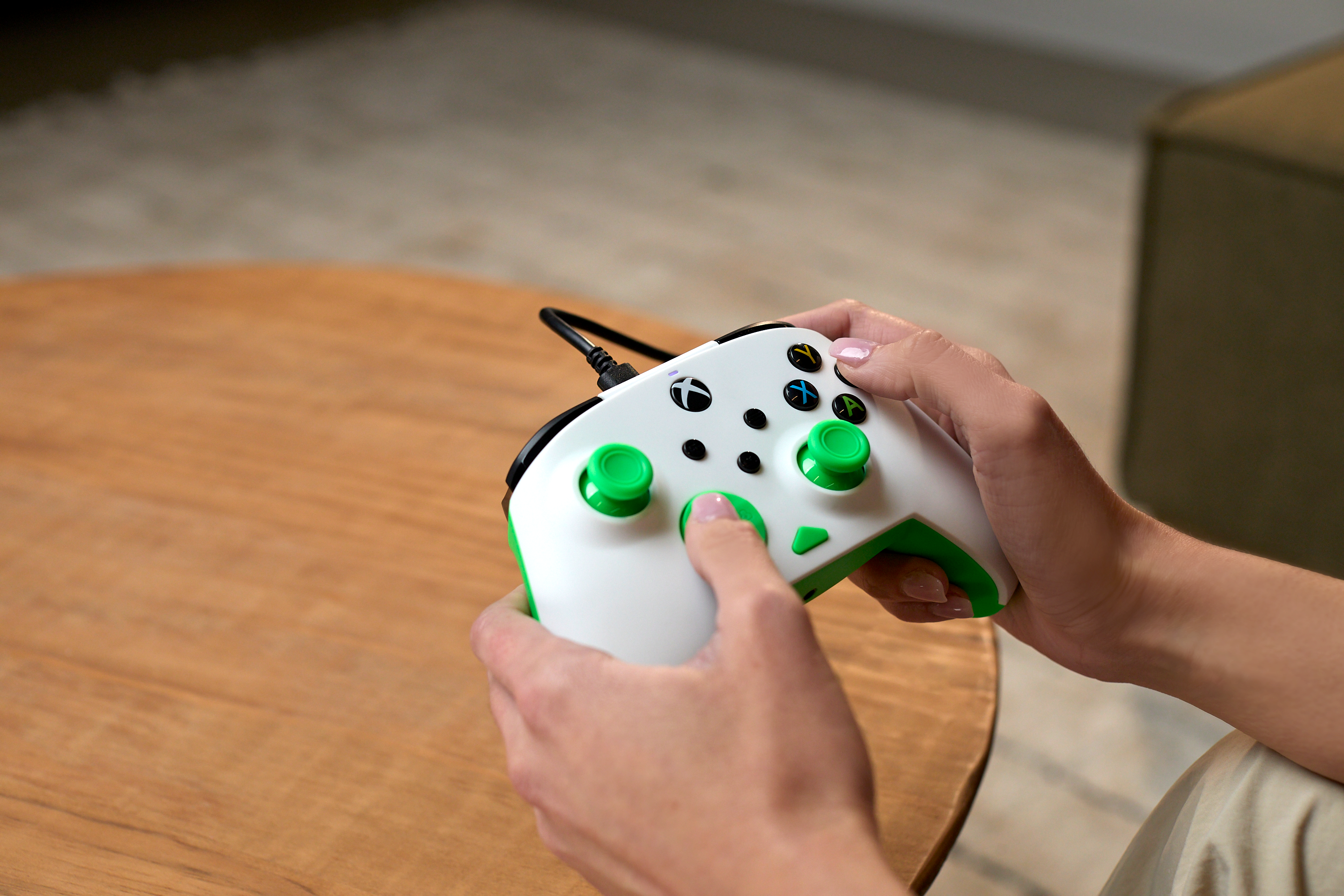 Osoba trzyma biało-zielony kontroler Xbox nad drewnianym stółem, skupiając się na dłoniach i przyciskach.