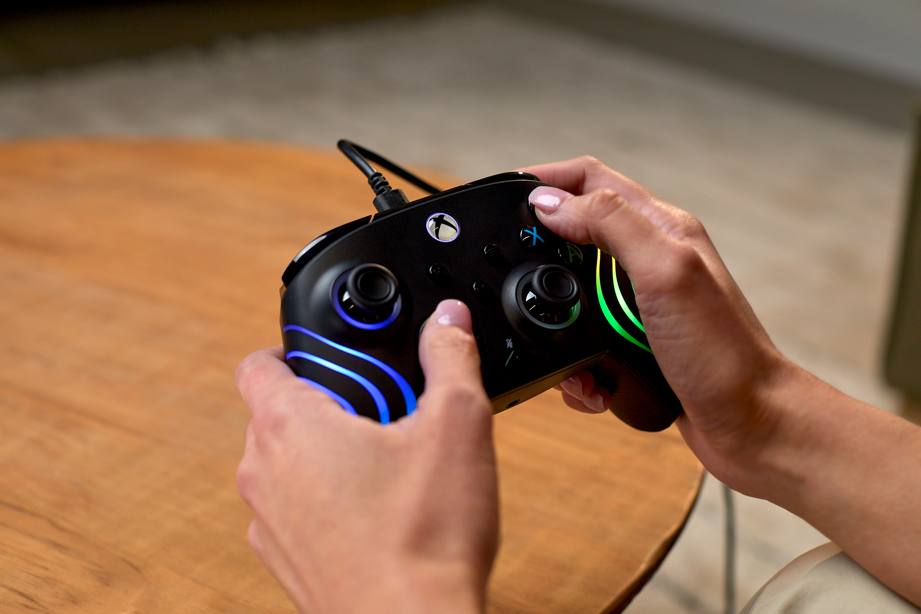 Dłonie trzymające czarny kontroler Xbox z niebieskimi i zielonymi diodami LED nad drewnianym stołem.