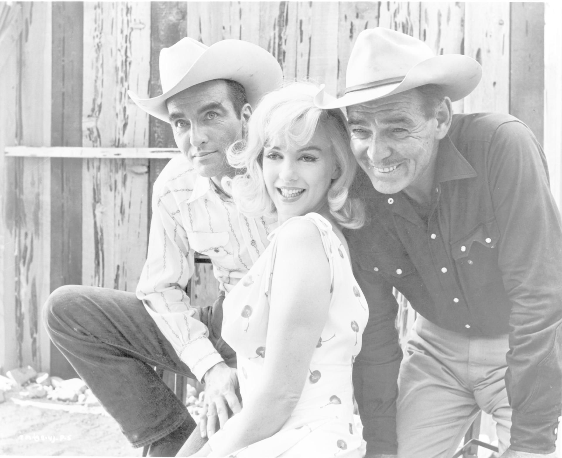 Marilyn Monroe sitzt zwischen zwei Männern mit Cowboyhüten und lächelt vor einer Holzwand.