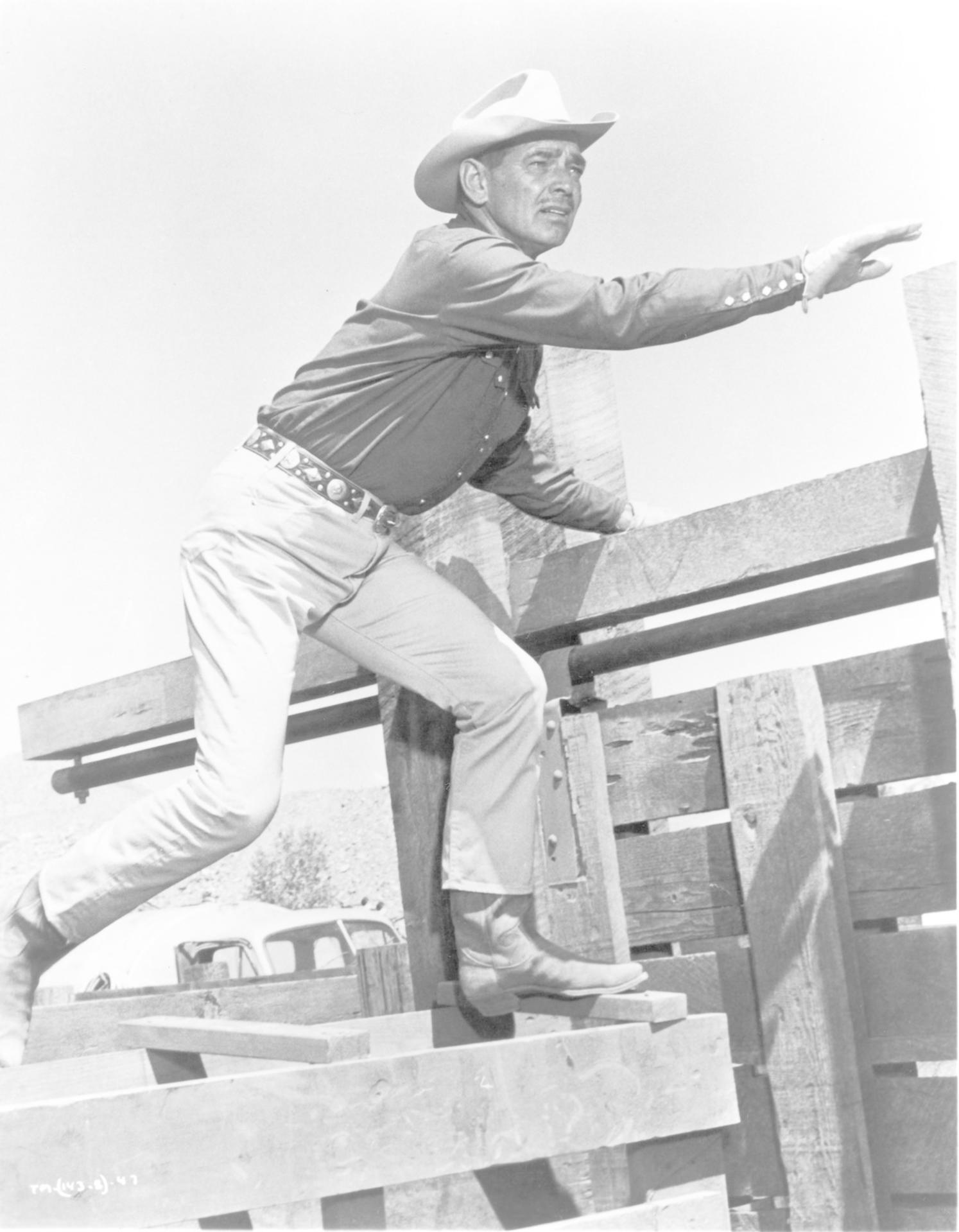 Ein Cowboy in Hut klettert über Holzzäune und zeigt mit der Hand nach vorne.