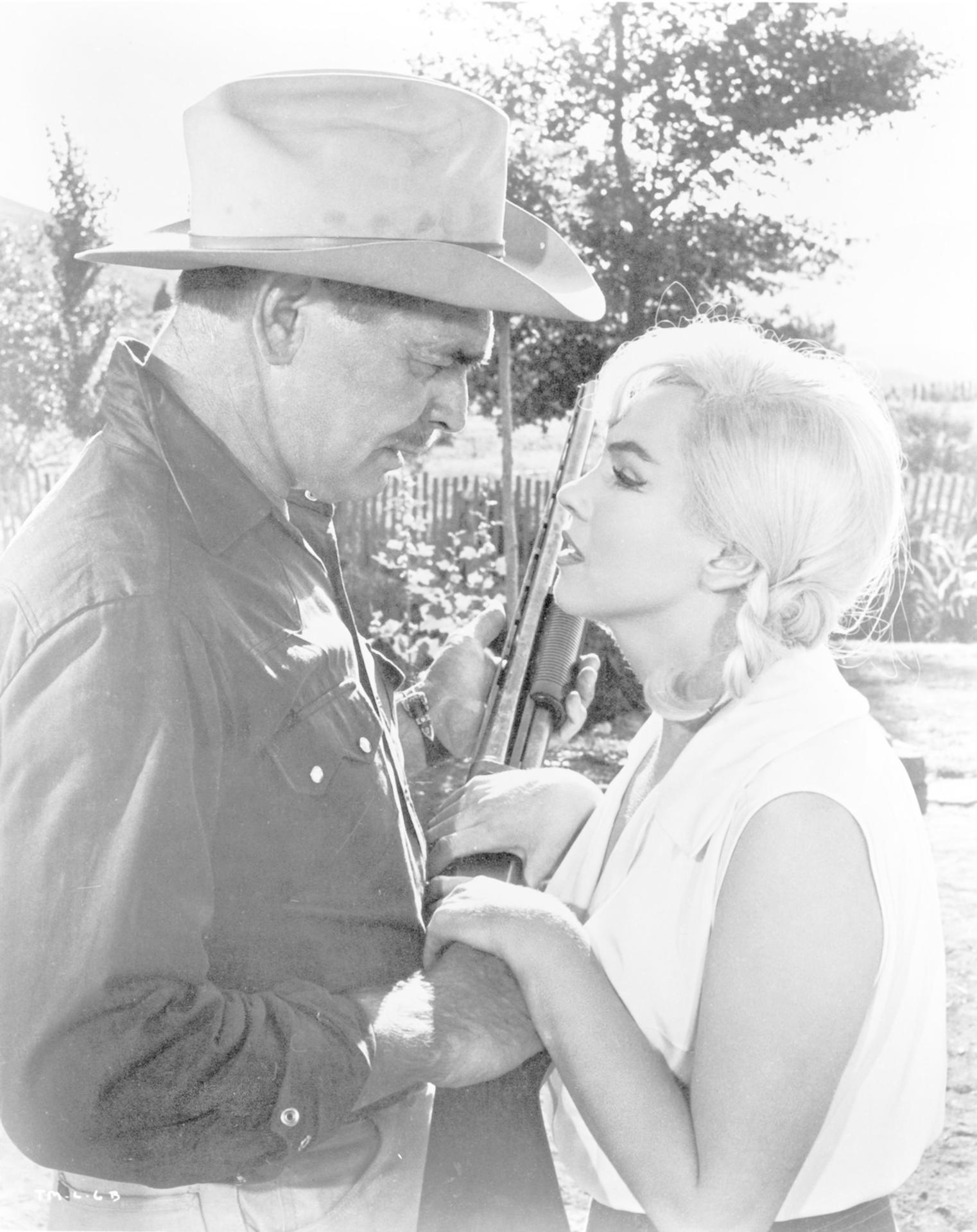 Ein Cowboy mit Hut und eine blonde Frau stehen sich draußen nahe gegenüber, eine Flinte ist zwischen ihnen.