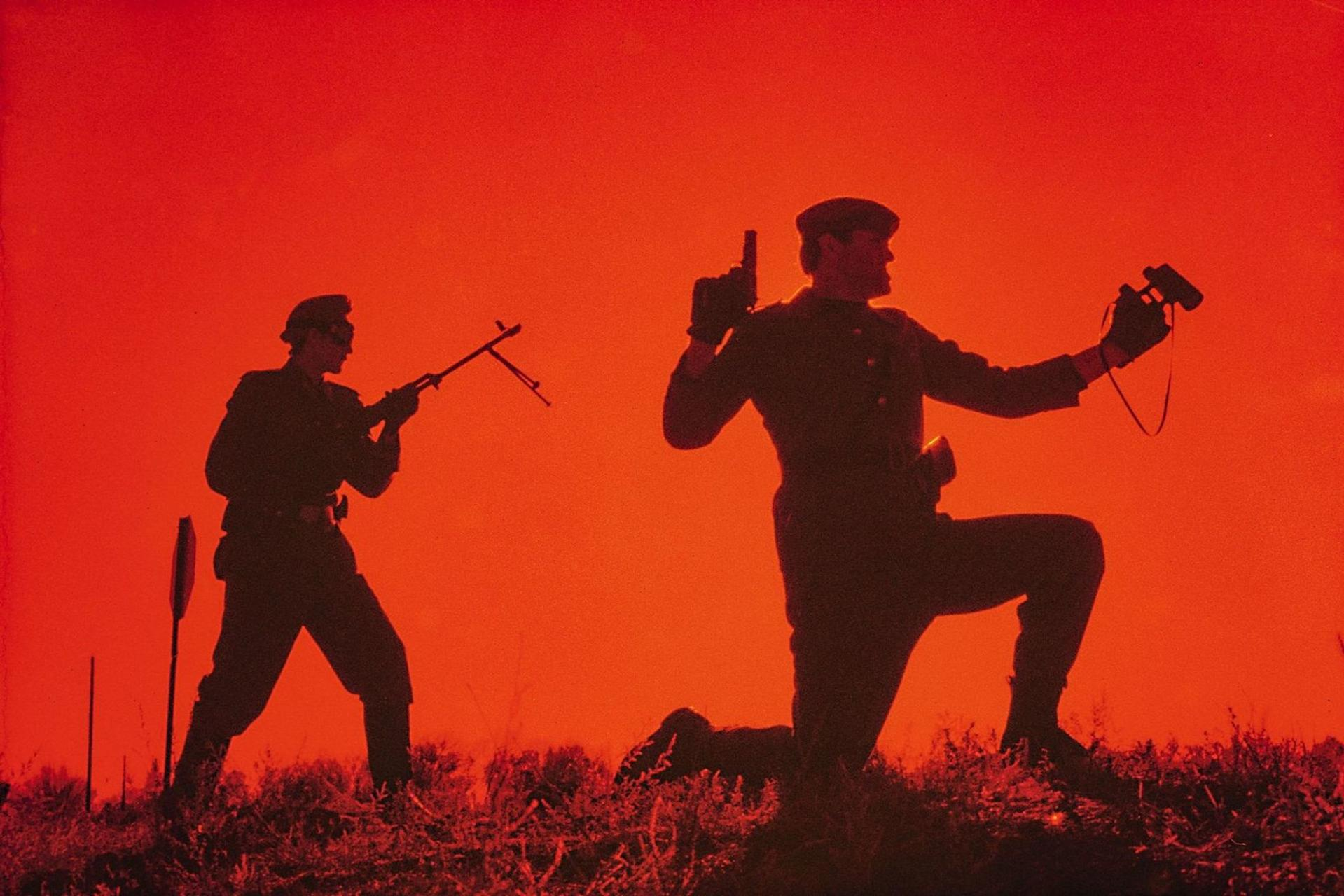 Zwei Soldaten im Gegenlicht vor rotem Hintergrund, einer hält ein Gewehr, der andere eine Pistole und ein Fernglas.