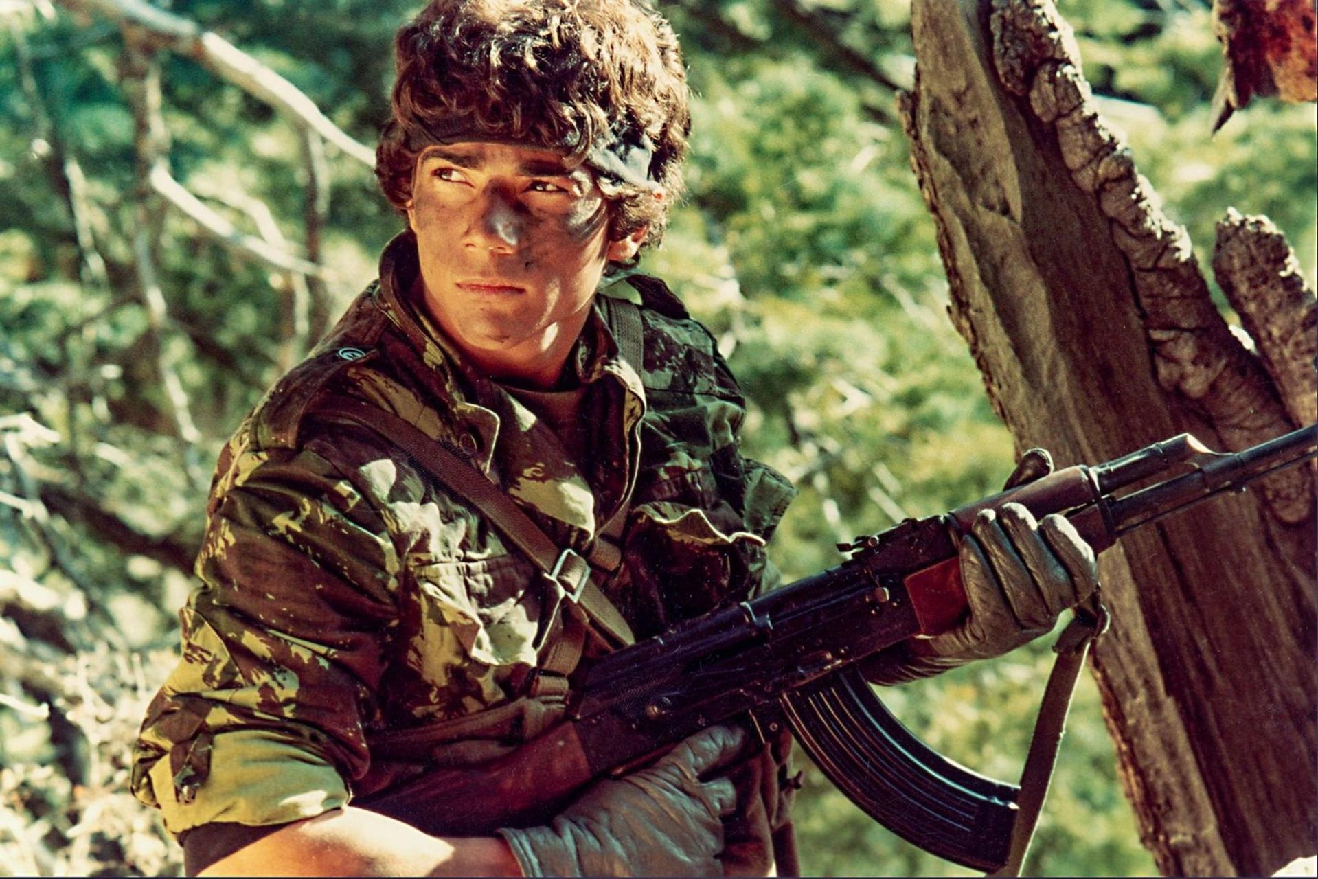 Ein Soldat mit Tarnkleidung und Stirnband hält im Wald aufmerksam eine Waffe.
