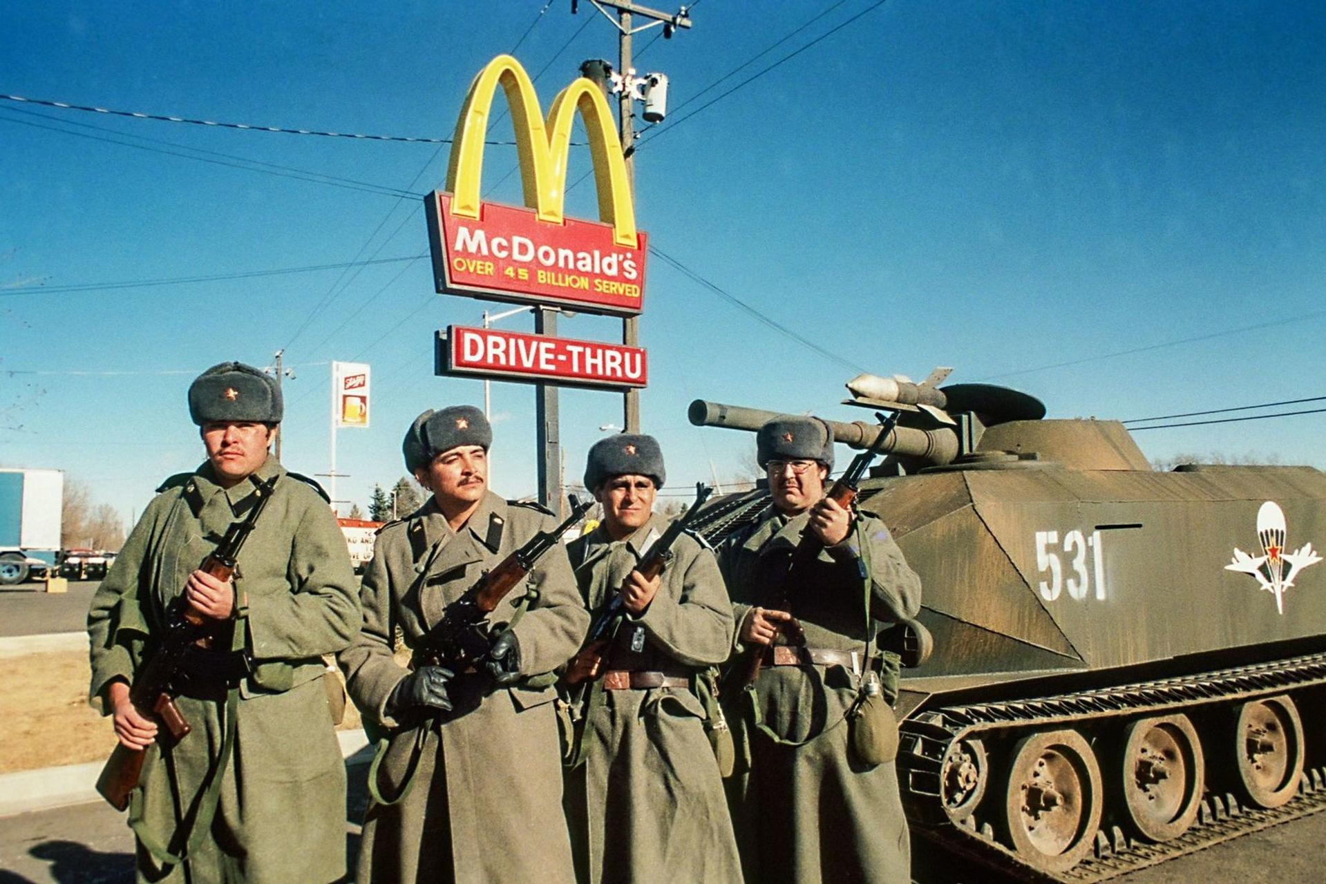 Vier Soldaten mit Gewehren und Pelzmützen stehen vor einem McDonald's-Schild und einem Panzer.
