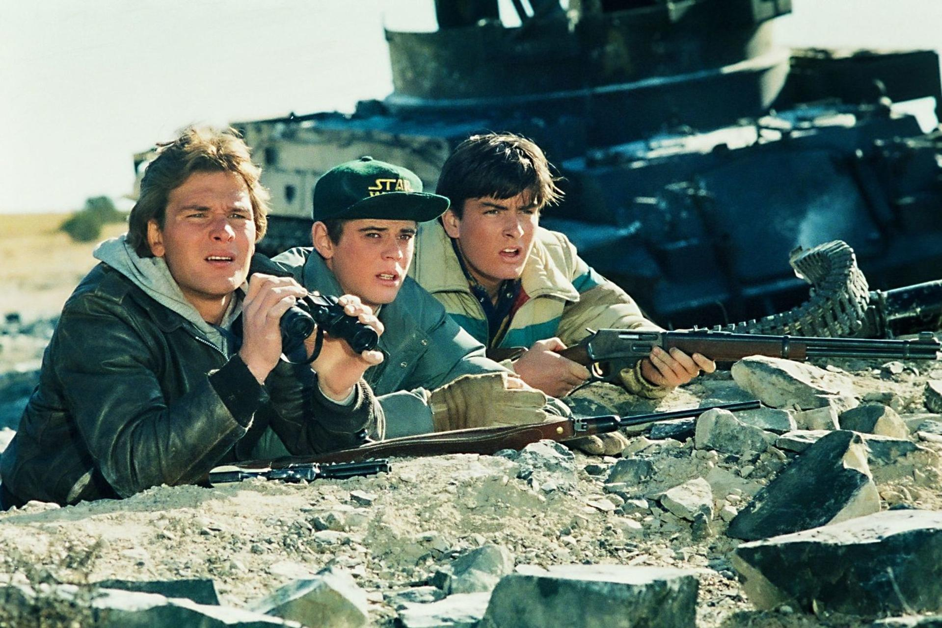 Drei junge Männer in einer Felslandschaft, einer mit Fernglas, einer mit Gewehr, im Hintergrund ein Militärfahrzeug.