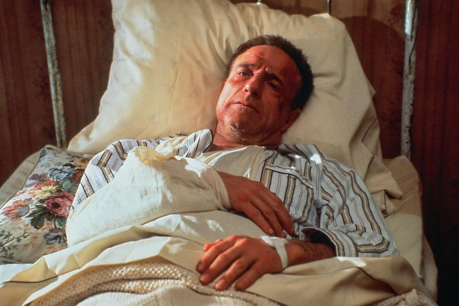 Ein verletzter Mann mit einem Gipsarm liegt im Bett und schaut besorgt nach oben.