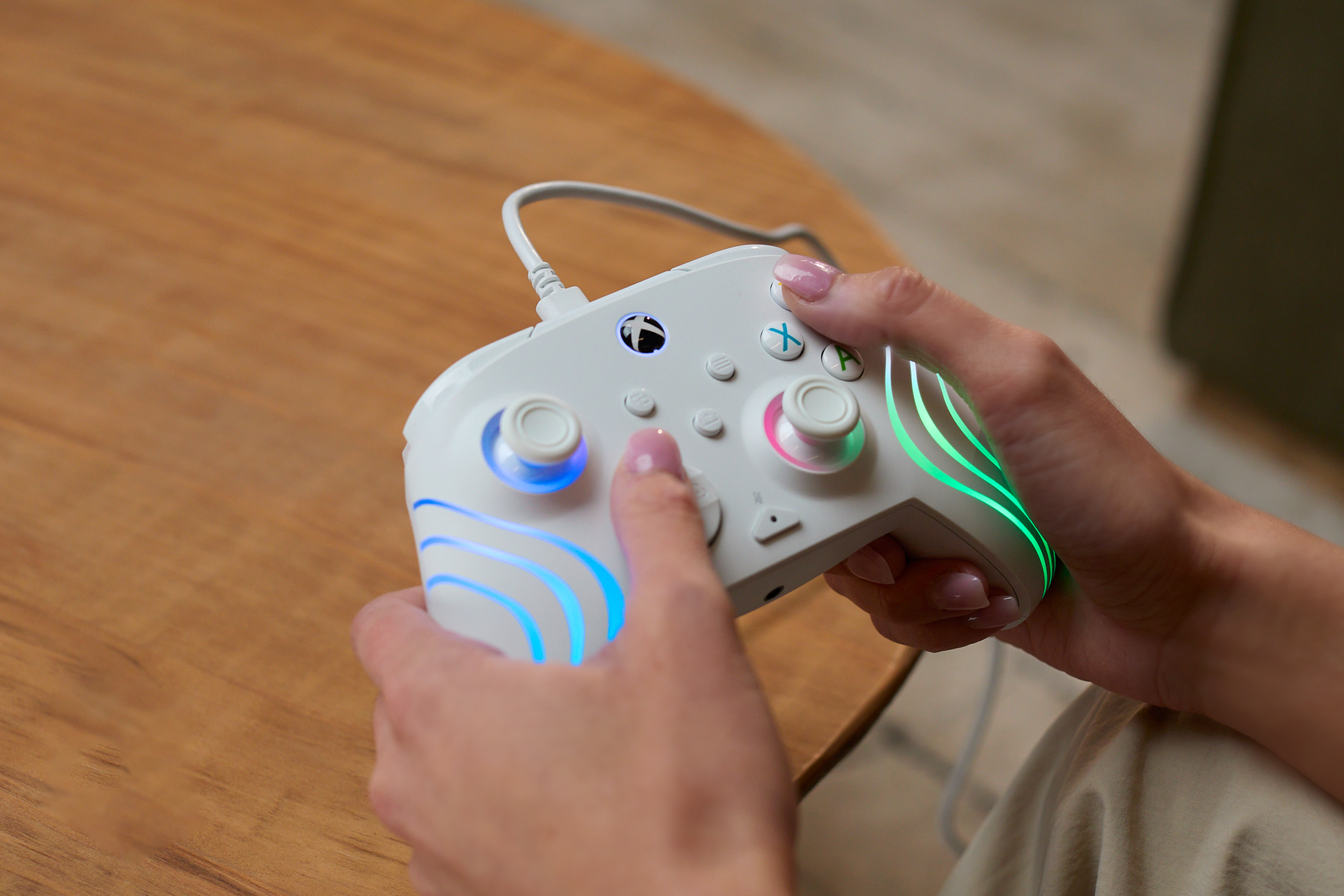 Une personne tient une manette Xbox blanche avec des LED colorées et un câble sur une table en bois.